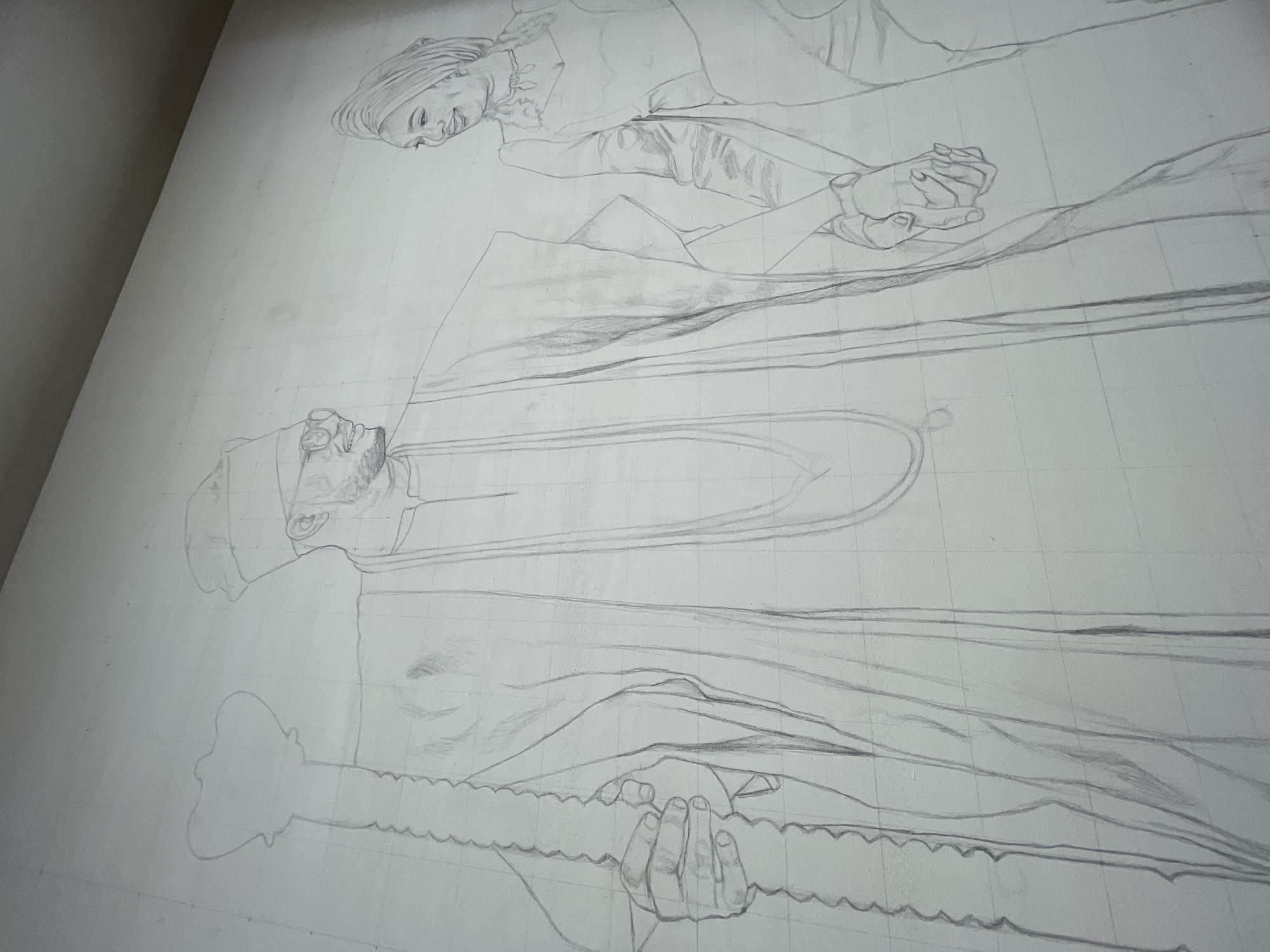 The image is a pencil sketch on a white surface, possibly a wall, marked with a grid pattern. Two human figures are depicted: one lying down at the top of the image and another below in a prone position. The figure at the top has a head covering and is smiling. Their hands are clasped together, and details of clothing and facial features are discernible.
Below, the second figure also appears to be lying down. They are wearing a cap or head covering. There is the suggestion of facial hair. The figure is holding an object that could be a staff or rod with an articulated surface.
The sketch appears to be a preparatory drawing for a larger artwork, with attention given to detail in the figures and their poses. The location in the city of Ado, Nigeria is not discernible from the visual information provided.