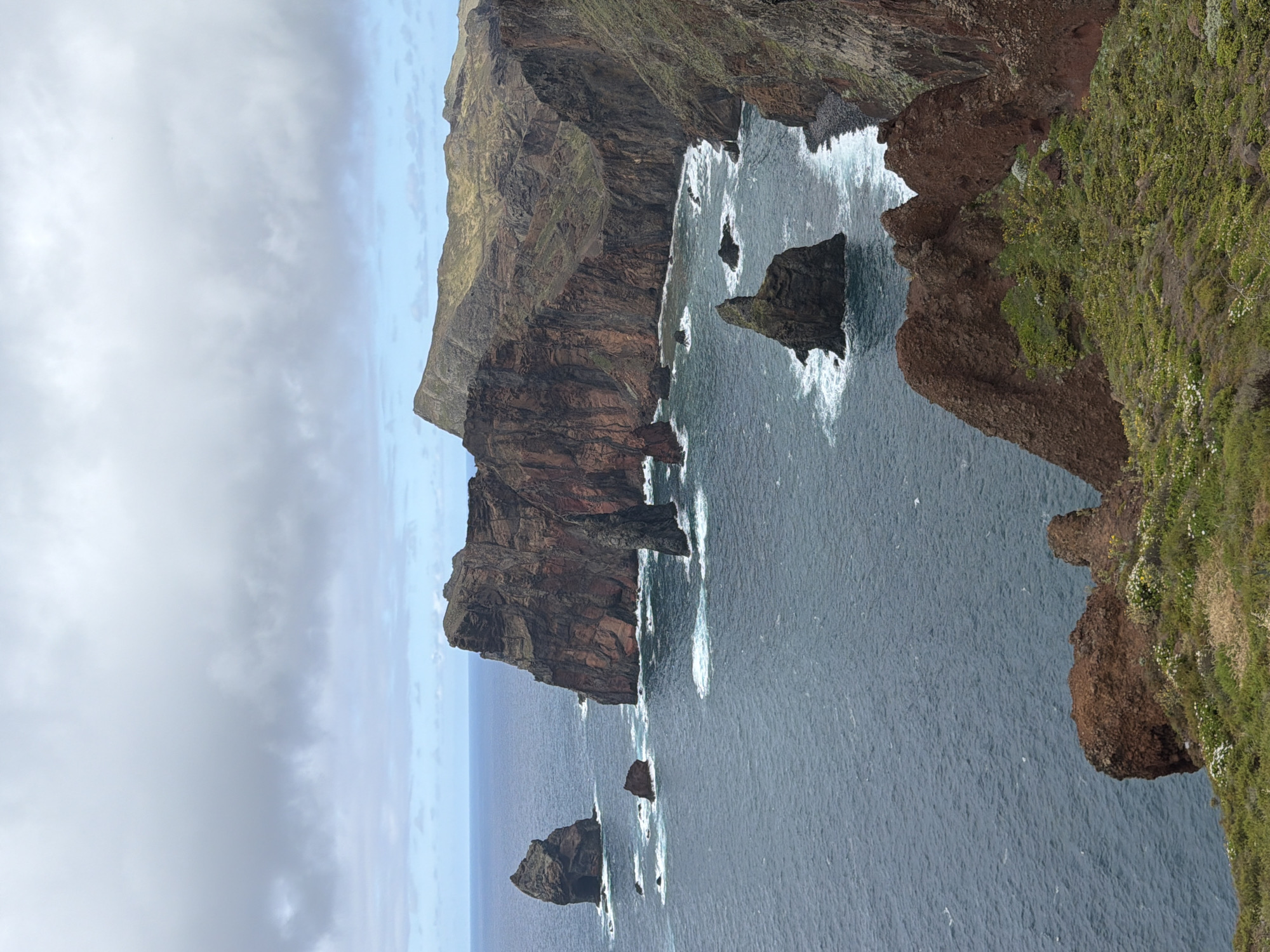 The image shows a coastal scene in Machico, Portugal. Rugged, reddish-brown cliffs rise dramatically from the dark blue ocean. Waves crash against the base of the cliffs and surrounding rock formations, creating white foam. Green vegetation covers parts of the cliff tops. The sky is overcast with grey clouds.