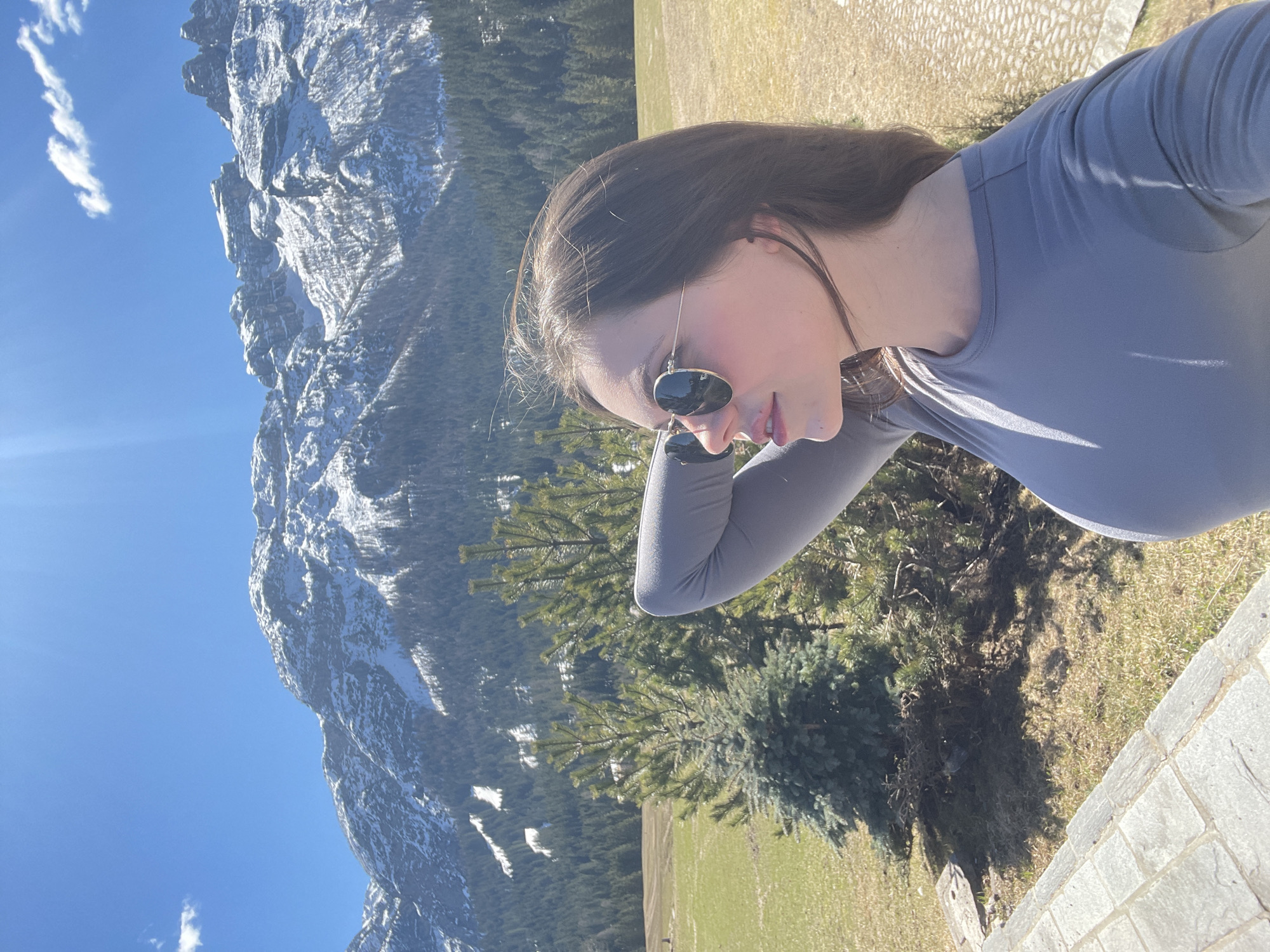 A woman wearing sunglasses and a grey long-sleeved shirt is taking a selfie. The background features a mountain with snow near Sappada, Italy, fir trees, and a blue sky with some clouds. A grassy area and a stone structure can also be seen.
