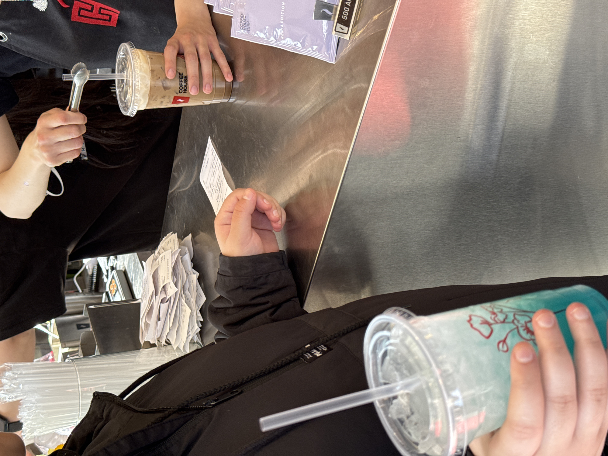 A close-up, tilted, high-angle shot depicts a transaction at a coffee shop, possibly located in Yerevan, Armenia. The foreground features a person in a black jacket holding a light blue drink in a clear plastic cup with a straw. Next to the drink, a stack of paper receipts is visible. In the midground, a person wearing a black shirt is preparing a coffee drink in a clear plastic cup, using tongs to stir it. The coffee cup bears the logo of "COFFEE HOUSE". The countertop is stainless steel and holds coffee packets. The background is indistinct, but suggests a cafe interior.