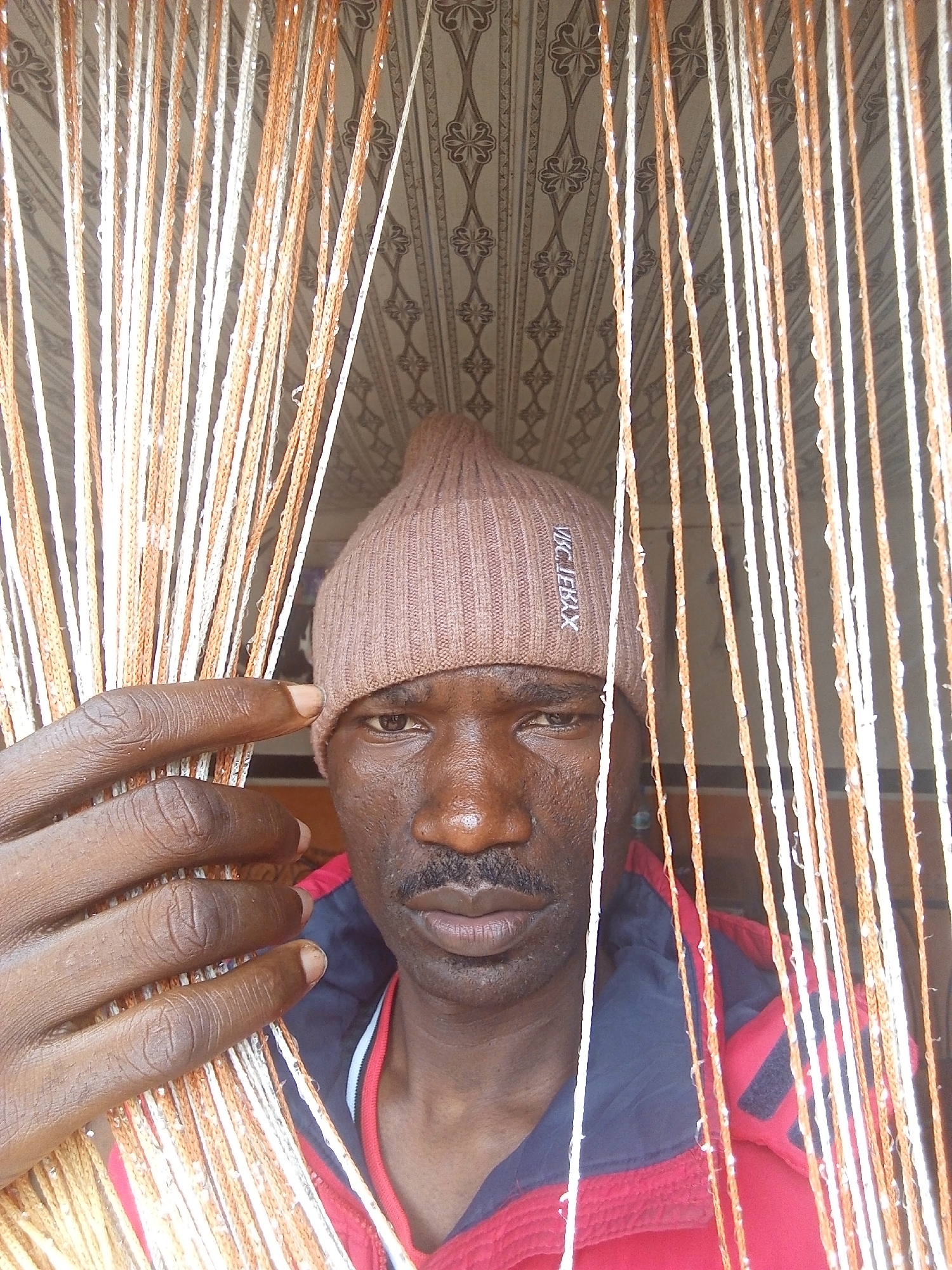 A medium, eye-level shot depicts a man wearing a brown knit cap, a red and black jacket with a red collar, and positioned behind a curtain of white and orange twisted strands. The location is suggested as Gwong, Nigeria. The man's left hand is visible, partially obscuring his face as he holds the curtain. Behind the curtain and above the man, a patterned ceiling is visible. The man is dark-skinned and has a mustache. The knit cap has lettering on it that is partially obscured. The environment is indoors.
