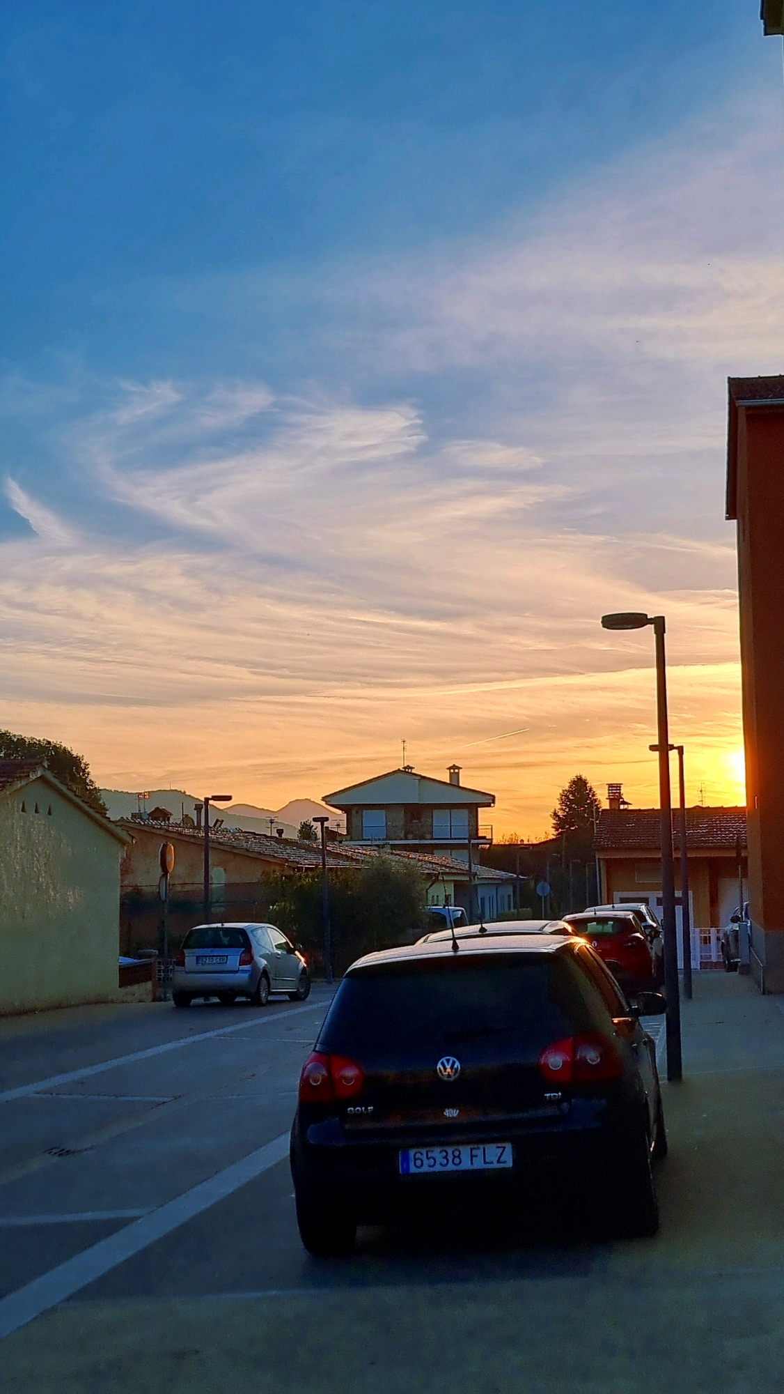 This is a vertical, eye-level, full-shot photograph taken at dusk in Sant Joan Les Fonts, Spain. The image captures a street scene with a warm, orange sunset illuminating the sky, creating a contrast with the deep blue of the upper atmosphere. Thin, wispy clouds stretch across the sky, painted with hues of pink and yellow from the setting sun.
In the foreground, a black Volkswagen Golf with the license plate "6538 FLZ" is parked facing away from the camera, angled slightly to the left. Its taillights are illuminated. To its left, a silver car is also parked, facing the same direction. Further down the street, other vehicles are visible.
The background features a residential area with a variety of buildings. A light-colored, stucco-walled house is on the far left, with a sloped, tiled roof. To its right, other houses with tiled roofs are visible, some with dormer windows. In the distance, rolling hills or mountains form a silhouette against the colorful sky. Streetlights line the right side of the road.
The overall atmosphere is serene and peaceful, typical of a quiet evening in a Spanish town. The light is soft and diffused, casting long shadows on the ground. There are no people visible in the frame. The image appears to be captured in late autumn or early spring, given the light and the color of the sky.