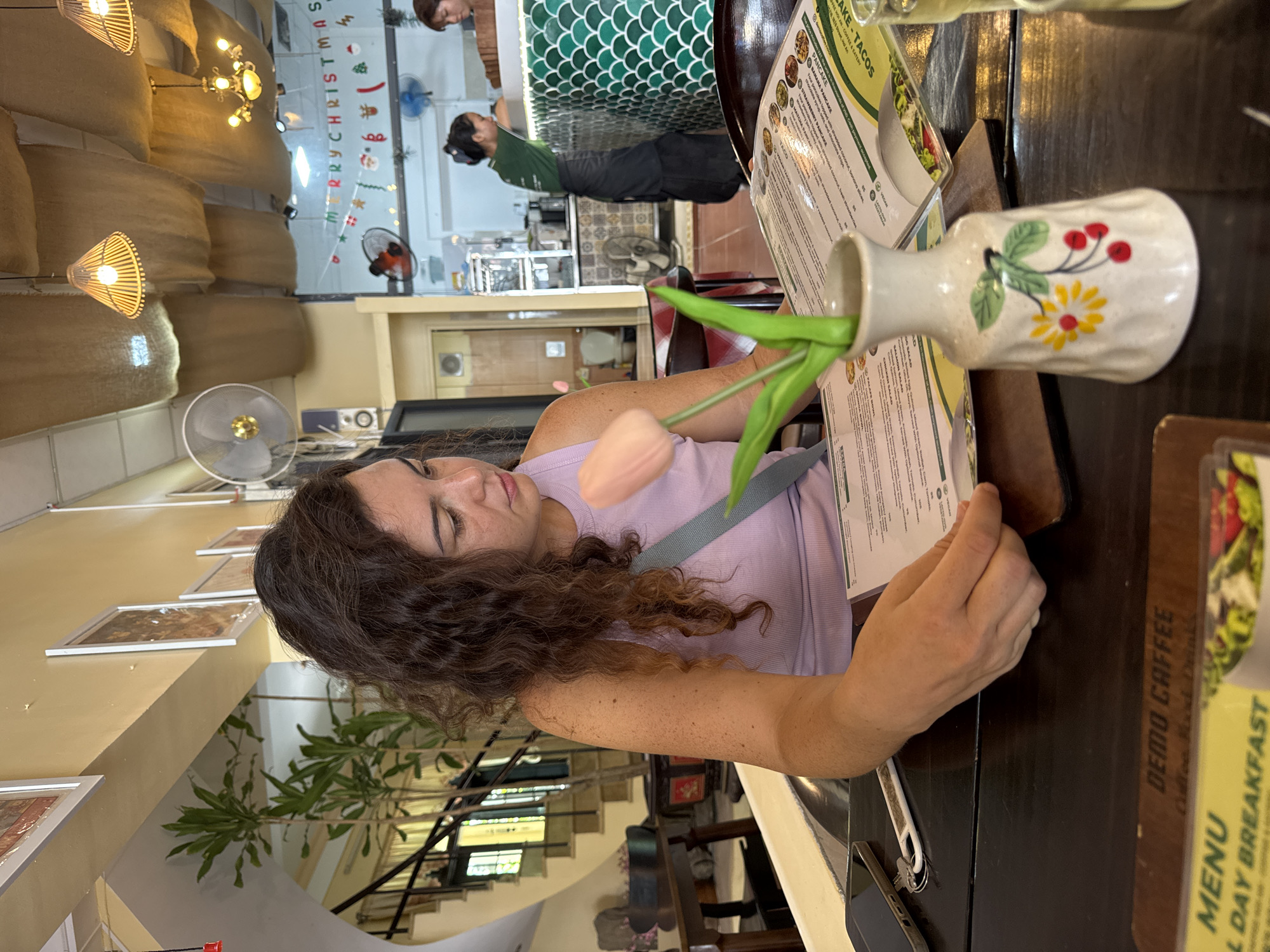 A young woman with curly brown hair is seated at a dark wooden table in a restaurant, looking down at a menu. She is wearing a light purple tank top and has a light blue strap across her chest. In front of her, on the table, are the menu, a smartphone, a set of keys, and a small vase with a single pink tulip and green leaves. The vase is decorated with painted flowers and cherries.
The restaurant appears to be decorated for Christmas, with a "Merry Christmas" banner visible in the background and festive lights. Behind the woman, another woman, likely a server, is seen in the background at a counter. The restaurant has a tropical feel with plants visible in the upper left corner and a staircase. The overall lighting is warm and inviting, with decorative lamps hanging from the ceiling, covered with burlap-like material. A portion of a glass with a light-colored beverage is visible on the right edge of the frame. The woman is in the process of ordering or deciding what to eat, as indicated by her focus on the menu. The scene suggests a casual dining experience.