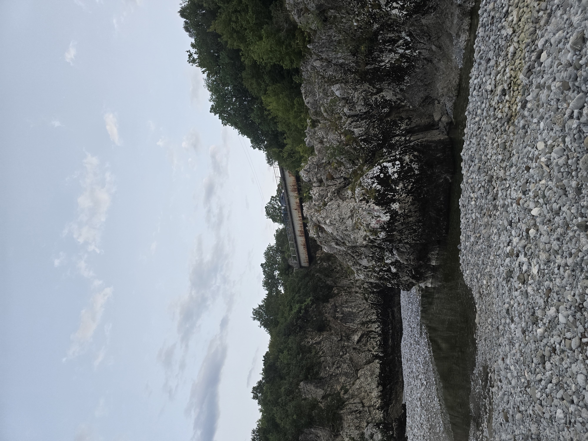 The image shows a rocky riverbed with a narrow channel of water. The riverbanks are steep and covered in rocks and dense green foliage. A rusty metal bridge spans the river in the distance, with a car visible crossing it. The sky is overcast with scattered clouds. The scene appears to be a natural landscape within the city of Bioče, Montenegro, characterized by rugged terrain and a flowing river.