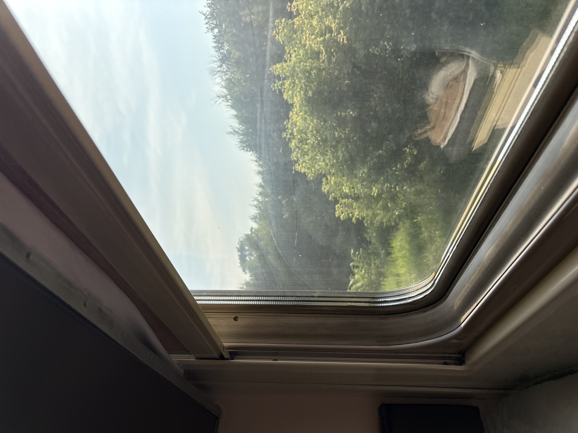 The image is an interior view from a train window looking out at a landscape. The window frame is metallic and beige. Outside, lush green trees and foliage are visible under a pale blue sky with scattered clouds. Sunlight is filtering through the leaves, creating dappled light. In the foreground, inside the train car, a dark material, possibly a curtain or shade, is visible along the top left of the frame. The overall scene suggests a journey through a rural or wooded area on a clear day. There is no visible indication of the city of Sovkhoznyi, Russia.