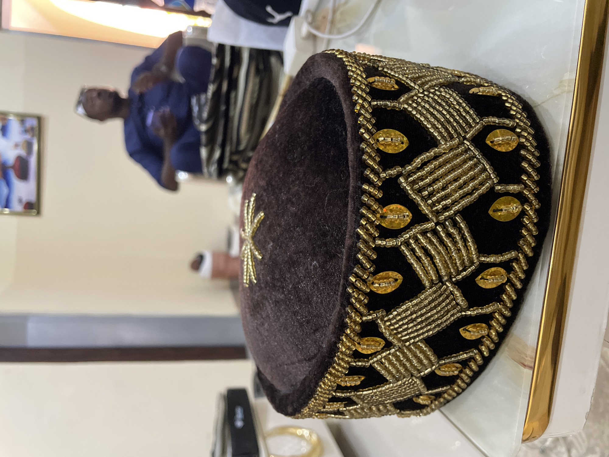 A traditional Nigerian hat, known as a fila, sits on a surface. The fila is brown with intricate gold beaded embroidery. In the blurred background, a man wearing a blue shirt stands in what appears to be an indoor setting. The specific location within Bar Jirgi Summa, Nigeria, cannot be determined from the image.