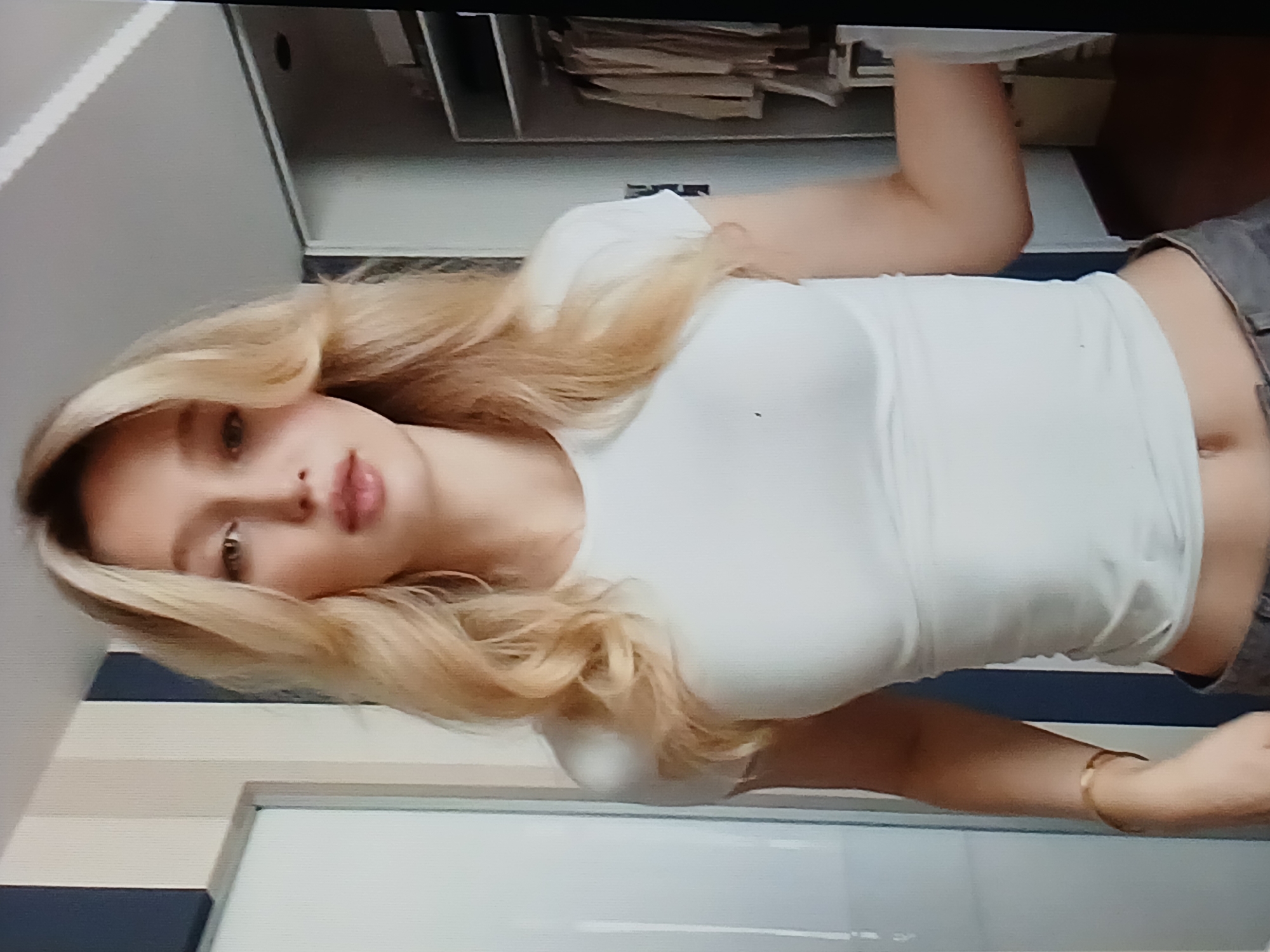This is a close-up, portrait-oriented photograph of a young woman with blonde hair, lying down. She is wearing a white tank top and what appear to be denim shorts. Her gaze is directed towards the viewer, and she has a neutral expression. The background is blurred, but suggests an indoor setting with shelves and what might be clothing or documents on them. To the left of the woman, there are vertical blue and white stripes, possibly part of a wall or furniture. A thin gold bracelet is visible on her left wrist. The lighting appears to be artificial, suggesting it is indoors.