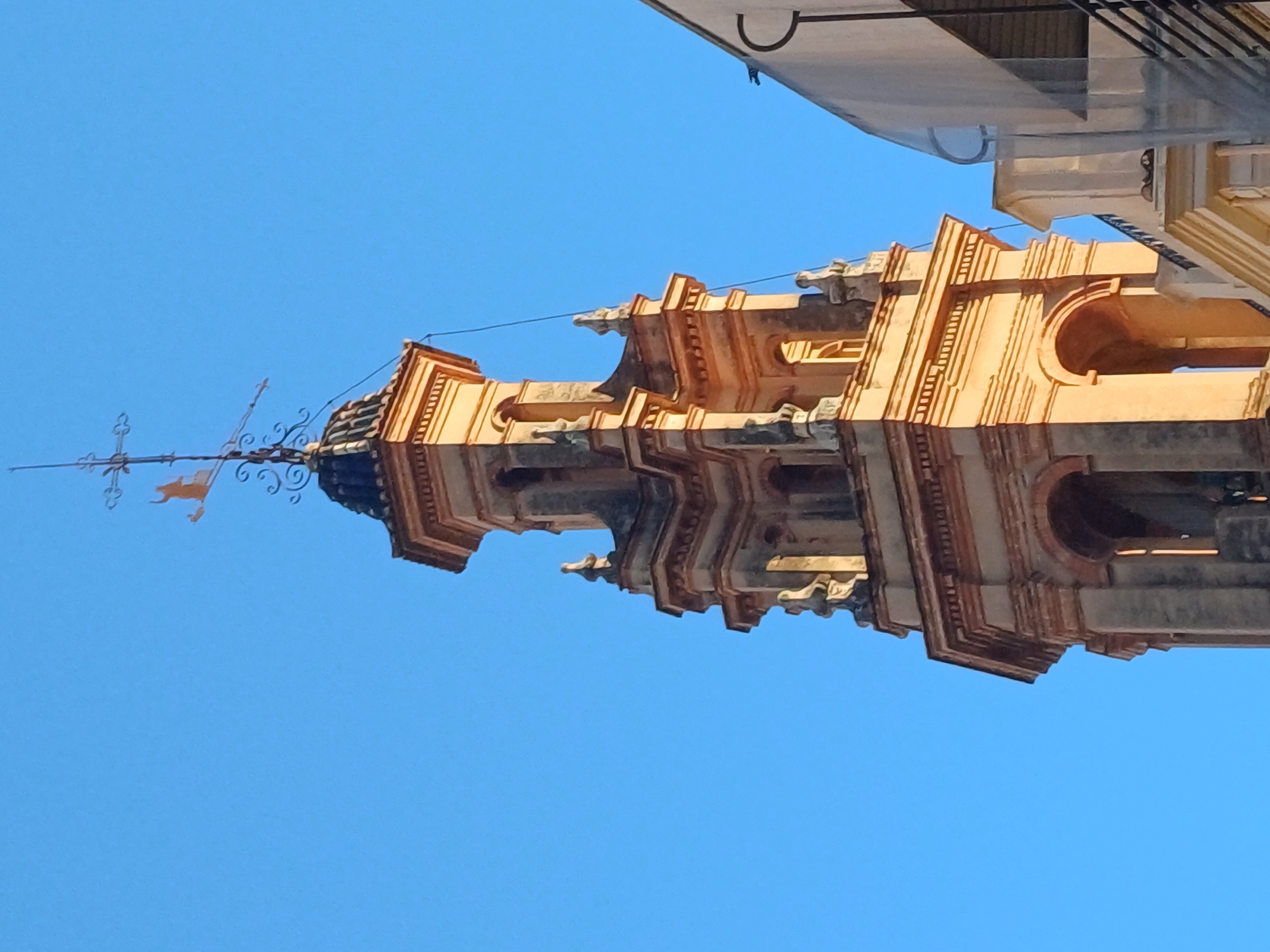 The image shows a low-angle shot of the top of the bell tower of Santa Catalina church in Valencia, Spain, against a clear blue sky. The tower is made of light-colored stone and has an intricate, multi-tiered design. A weather vane with decorative metalwork and a figure of a rooster is visible on top of the tower. The corner of a white building is visible in the upper right corner of the frame.