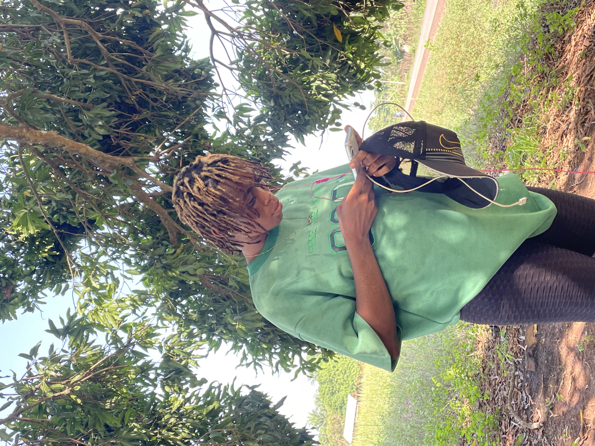 A person with light brown dreadlocks is standing outdoors in what appears to be a grassy area with trees. The person is wearing a loose-fitting, light green t-shirt, black leggings, and a gold chain around their neck. They are holding a black baseball cap with a gold Nike logo in their left hand and a mobile phone in their right hand, looking at the screen. A white charging cable is plugged into the phone. The background consists of trees and greenery, suggesting a natural or park-like setting. Given the prompt mentioning Ogbe, Nigeria, it is assumed that this scene takes place within the Ogbe area.