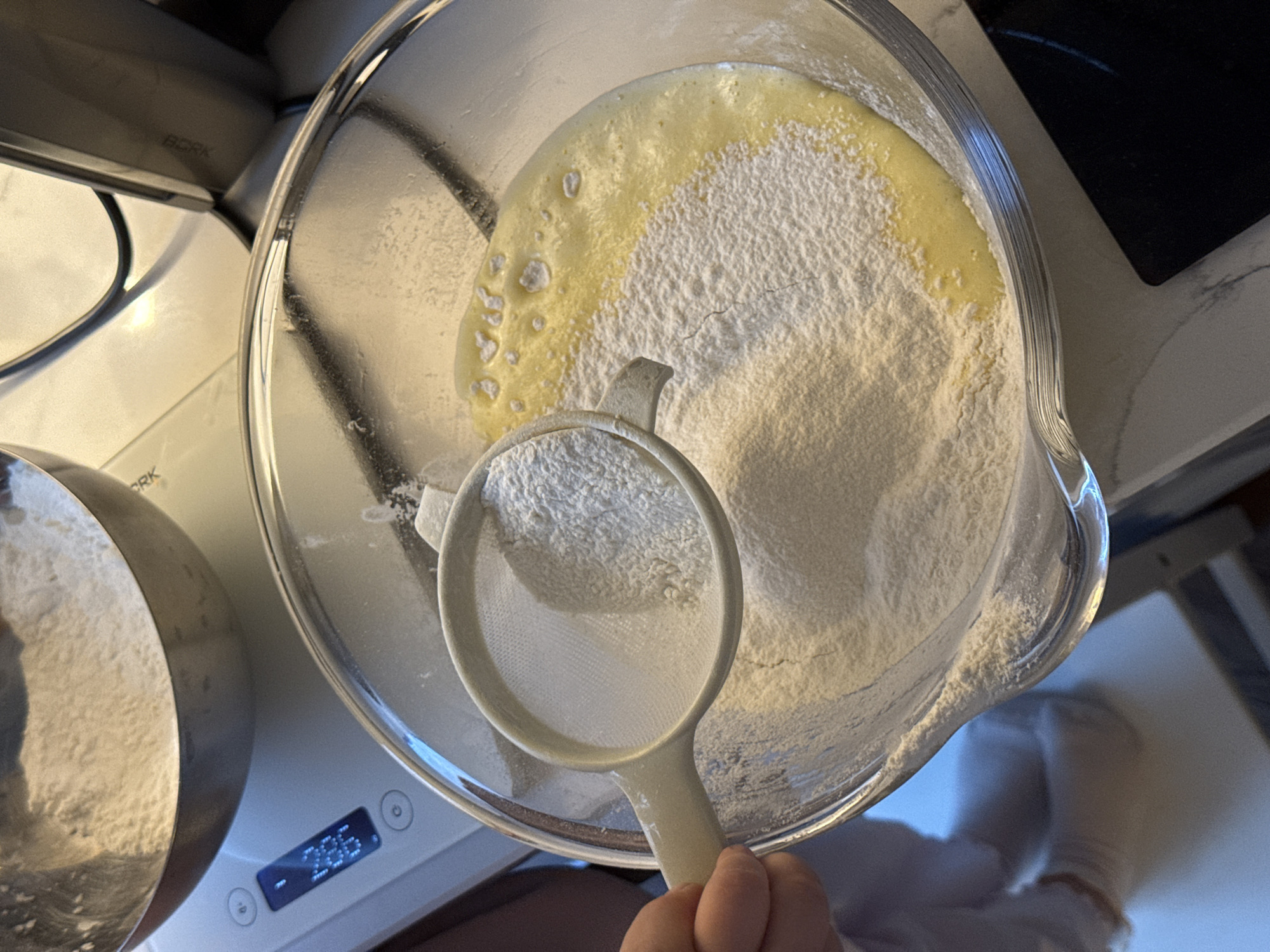 A top-down view captures a cooking scene indoors. A clear glass bowl is filled with a mixture of white powder and a yellowish liquid. A white plastic strainer is being held by a person out of the frame, containing more of the white powder above the bowl. To the left, a silver pot sits on a white digital scale, displaying "266" on the screen. Above the pot and scale, a silver appliance with the brand "BORK" visible sits on a kitchen counter. The background includes a dark stovetop on a white countertop. A gloved hand is visible at the bottom-right. The scene is most likely taking place in Moscow, Russia, given the presence of the "BORK" brand.