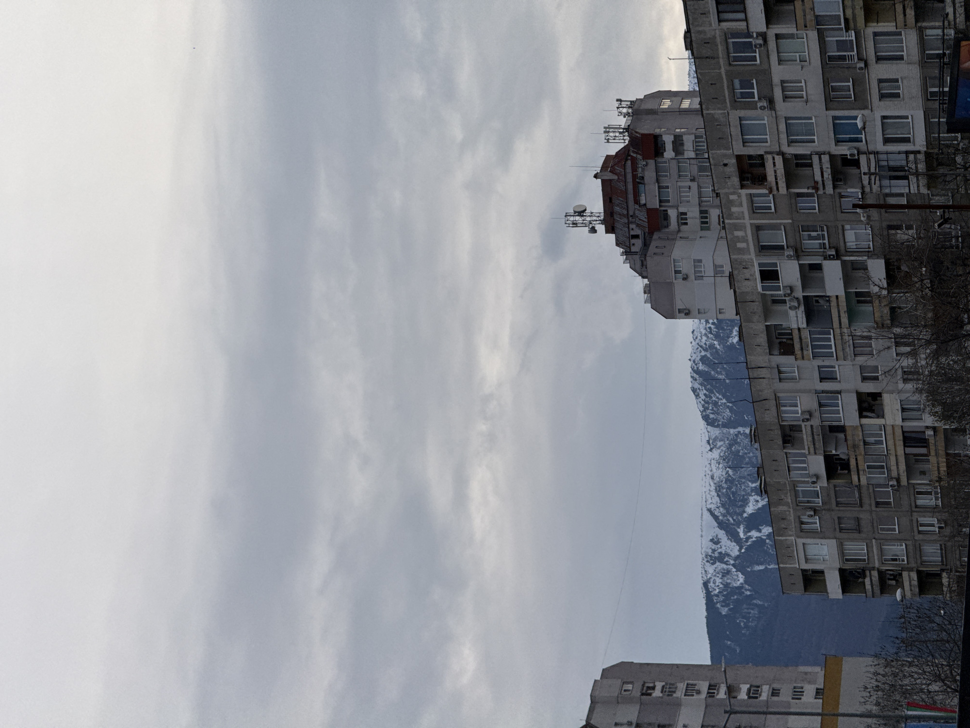 The image shows a cityscape with a cloudy sky. On the right side, there are several tall, concrete residential buildings with multiple windows and balconies. Some buildings have antennas on their roofs. In the background, partially obscured by the buildings, are snow-capped mountains. The location is likely in Sofia, Bulgaria, where similar architectural styles and mountainous backdrops are common. Bare tree branches are visible near the buildings. Another building, shorter than the rest, is seen near the bottom of the frame.