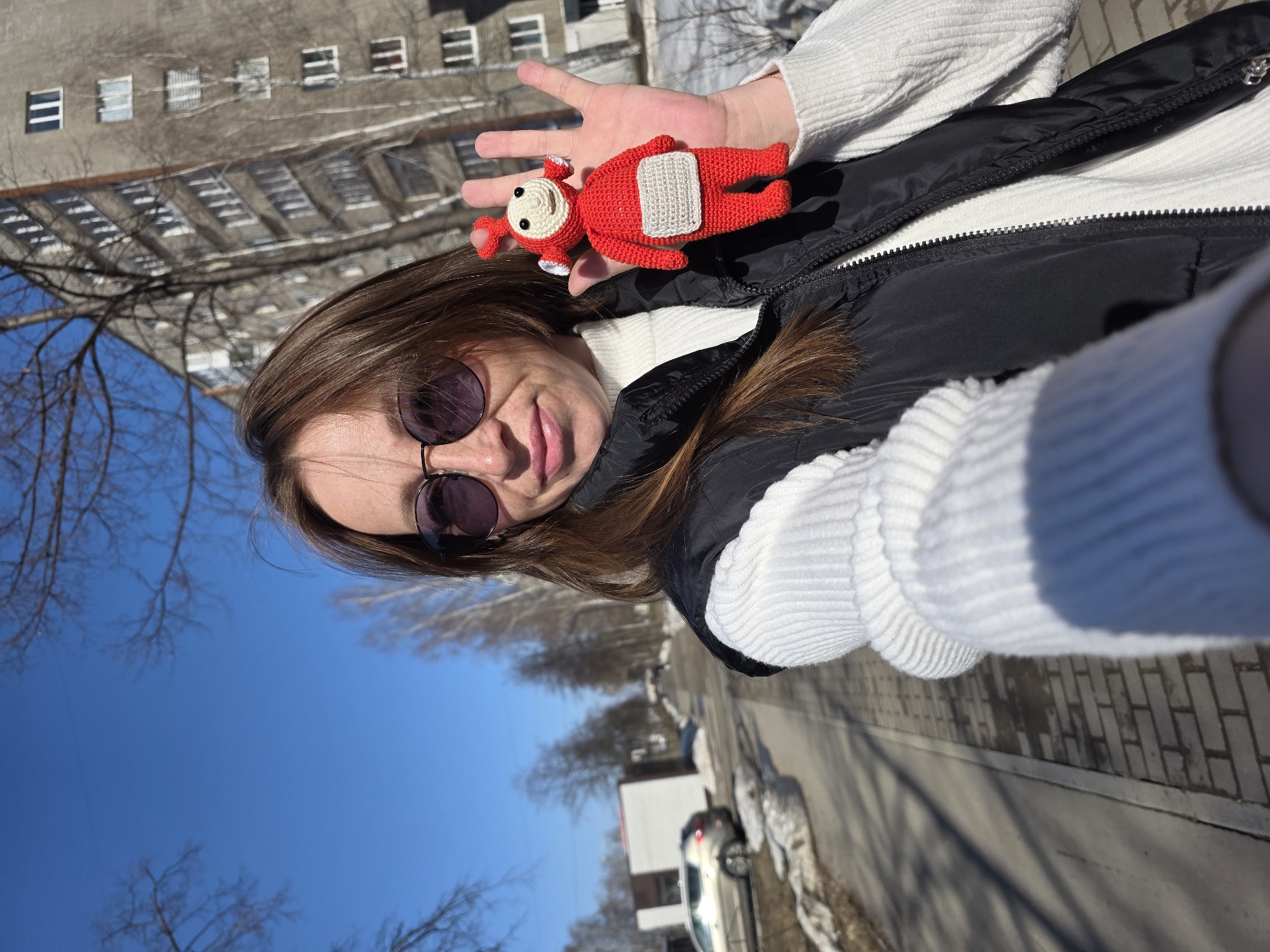 A woman is taking a selfie outdoors. She is wearing sunglasses, a white sweater, and a black vest. She is holding a red crocheted toy in her outstretched hand. The background shows a street, a building with many windows, bare trees, a blue sky, and a parked car. The image was taken in Chaikovsky, Russia.