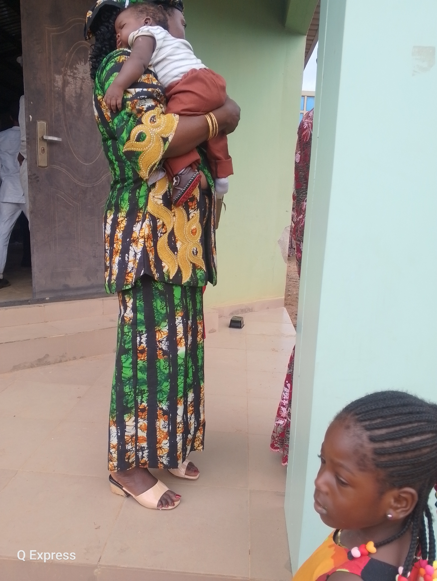 A woman in a vibrantly patterned green, black, and orange dress holds a sleeping baby. The baby is dressed in a white and striped shirt and brown pants. The woman's dress has intricate gold embellishments on the bodice. She is wearing tan heeled sandals. To the right, a young girl with braided hair is visible from the chest up. She is wearing a colorful dress with a beaded necklace. The scene appears to be outdoors, with a light green wall to the right and a building entrance with a brown door to the left. The ground is paved with light-colored tiles. The photograph is likely taken in Sabojibi, Nigeria, as indicated by the watermark "Q Express".