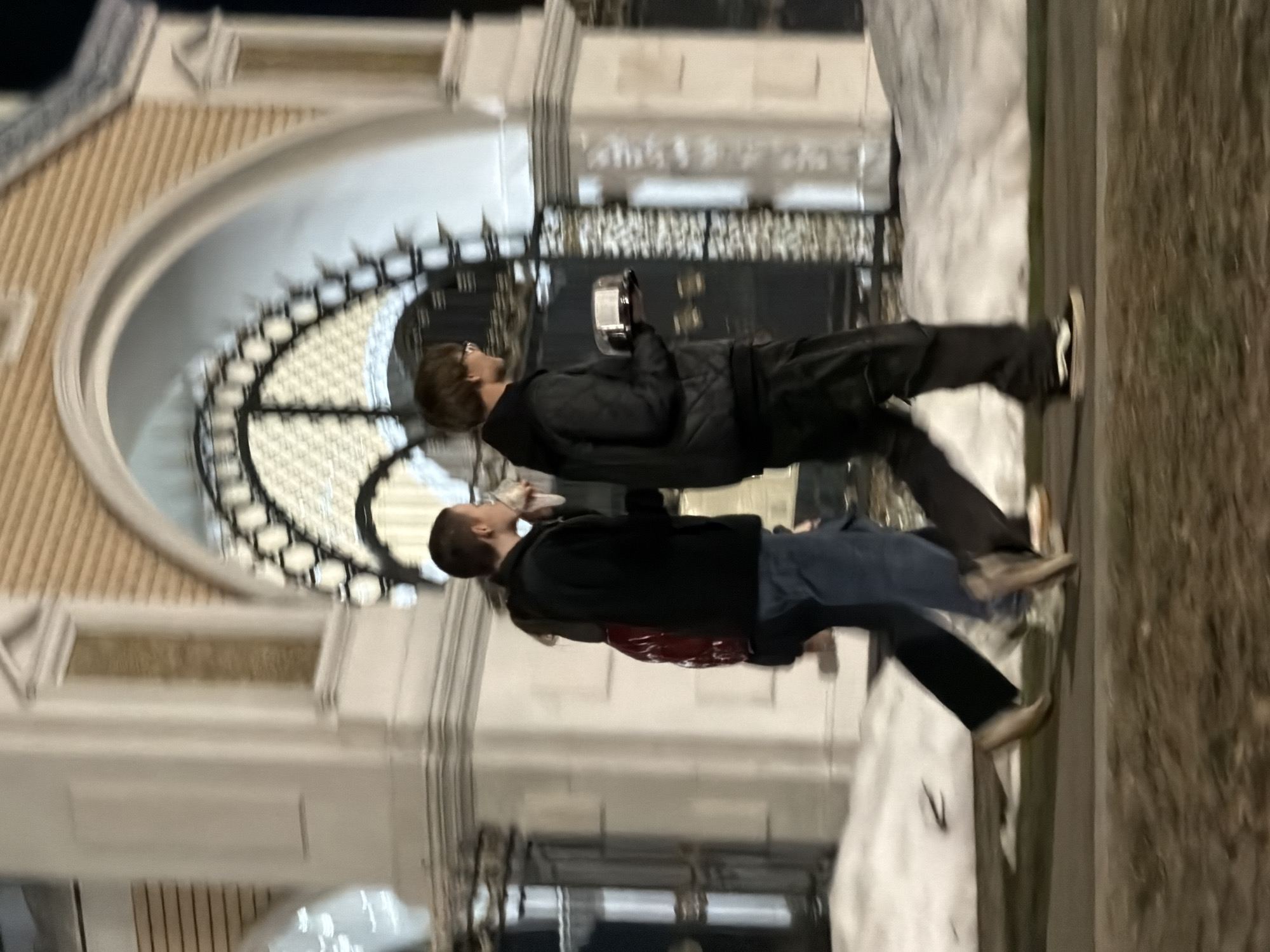 Two people stand outside a building featuring an arched entry with decorative ironwork. One individual holds a device, possibly a phone or camera, in their hand. The location is Izhevsk, Russia.
