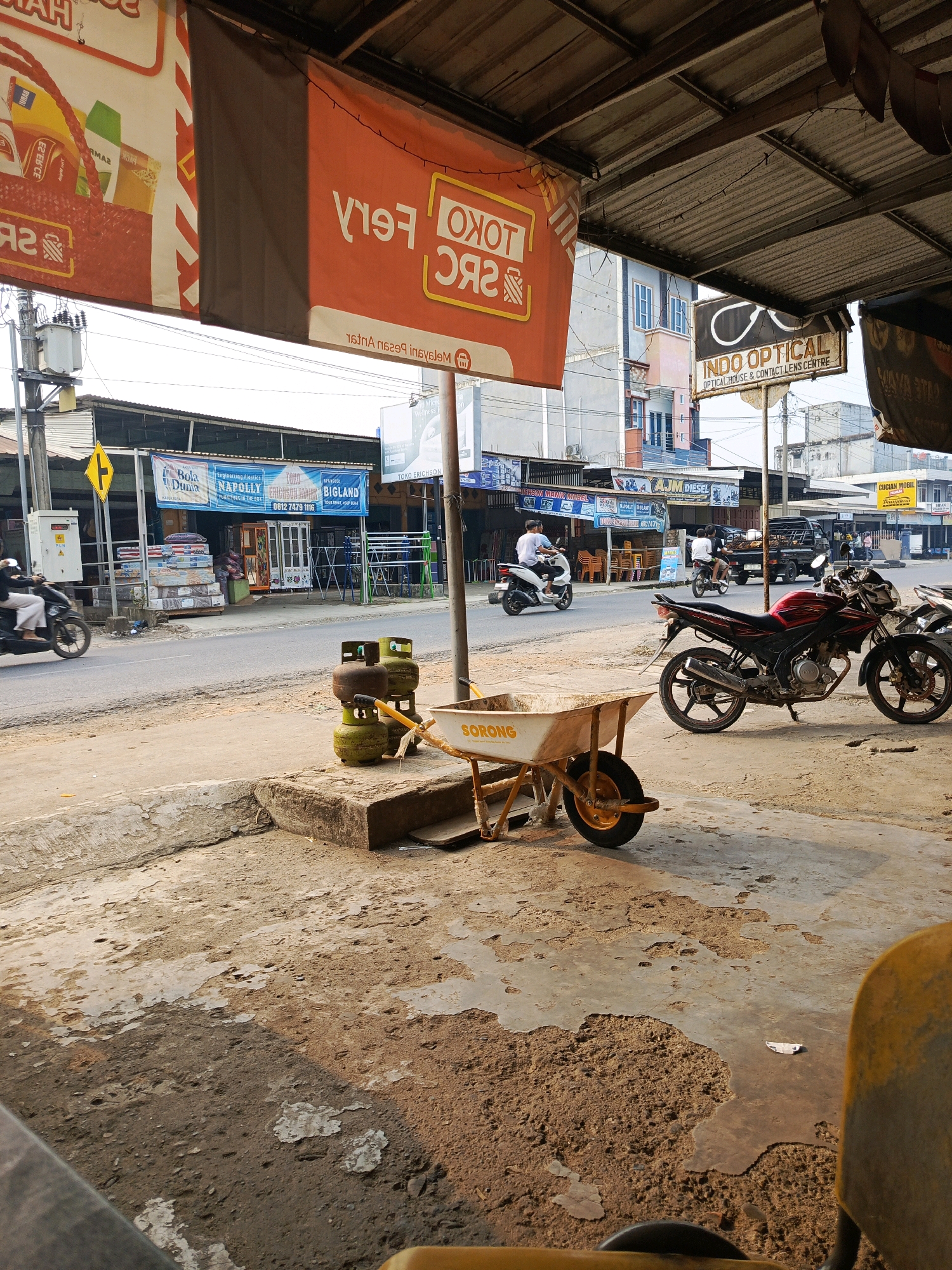 The image depicts a street scene in Megang Sakti II, Indonesia, under a corrugated metal awning. In the foreground, a worn concrete ground is visible with patches of dirt and debris. A yellow wheelbarrow with the brand "SORONG" is positioned on a raised concrete block. To the right, a stack of green and brown propane gas tanks rests on a similar block. A red motorcycle is parked on the right side of the street.
Across the street, a row of shops is visible. A banner with red and white stripes hangs on the left, and an orange banner with white text reads "TOKO FERRY". Further down the street, businesses include "Bola Dunna", "NAPOLLY Furniture in the Box", "TOKO ANGKASA", and "BIGLAND". A yellow diamond-shaped sign with a black arrow is attached to a utility pole. Two individuals on a motorcycle are seen on the left, and another motorcycle is ridden by two people in the middle ground. A black truck is parked further down the street. A sign for "INDO OPTICAL" with an eyeglasses logo is prominently displayed on a pole.
The overall scene suggests a commercial area with shops, street vendors, and typical street activity in Indonesia. The lighting indicates it is daytime, likely late afternoon given the long shadows.
