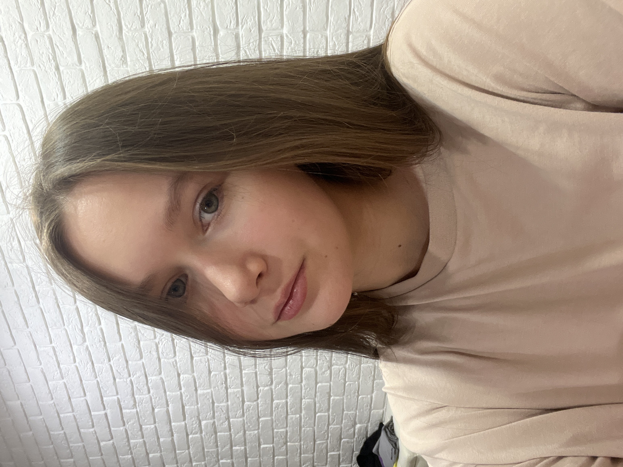 A young person with fair skin and shoulder-length light brown hair is positioned in front of a white brick wall. They are wearing a light pink t-shirt. The image appears to be taken indoors.
There is no location information in the city of Chaikovsky, Russia visible within the image.
