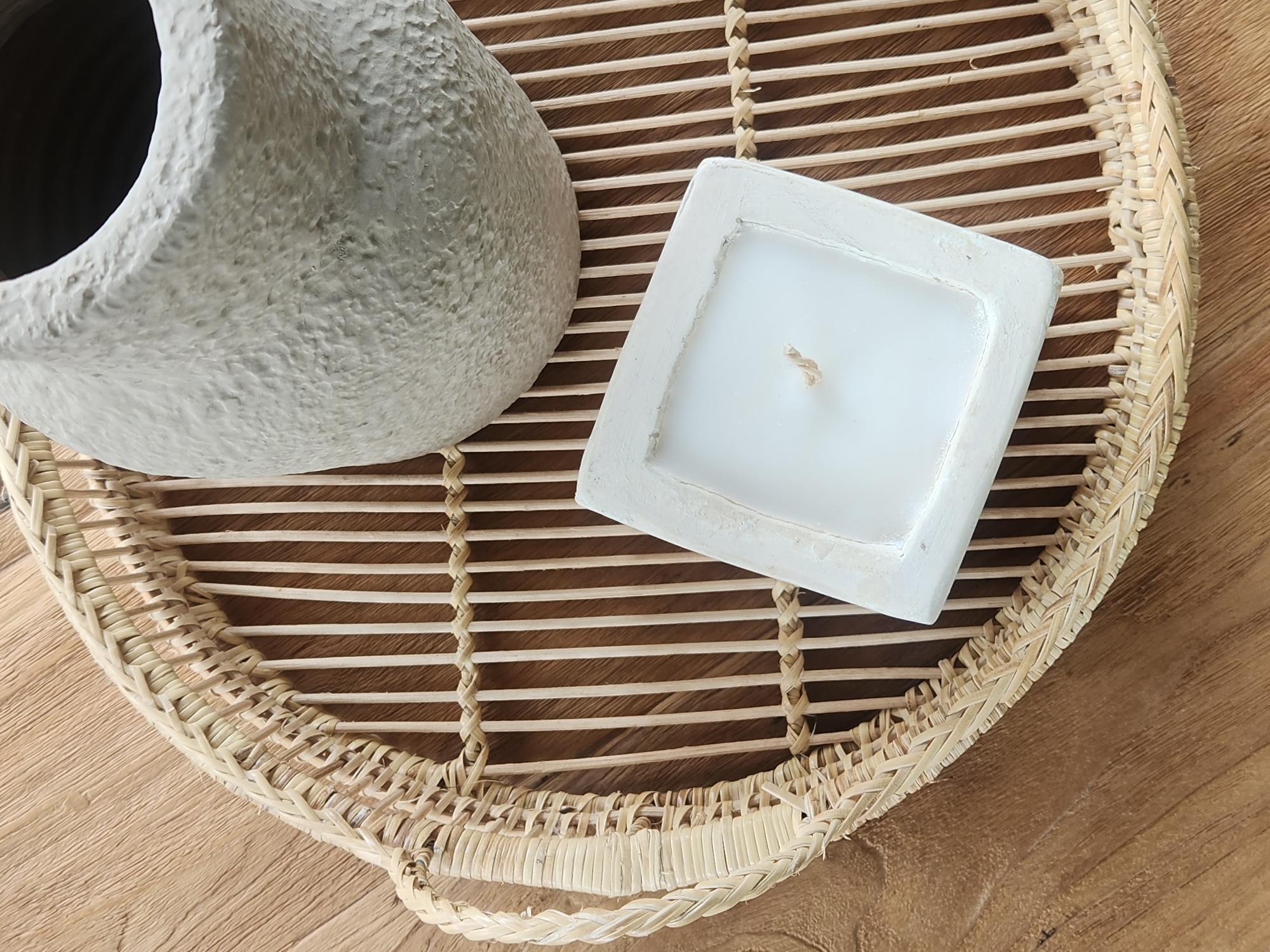 A top-down shot shows a woven wicker tray on a wooden surface. On the tray are a square white candle with a wick and a textured, light gray vase. The scene is indoors, and there are no people present.