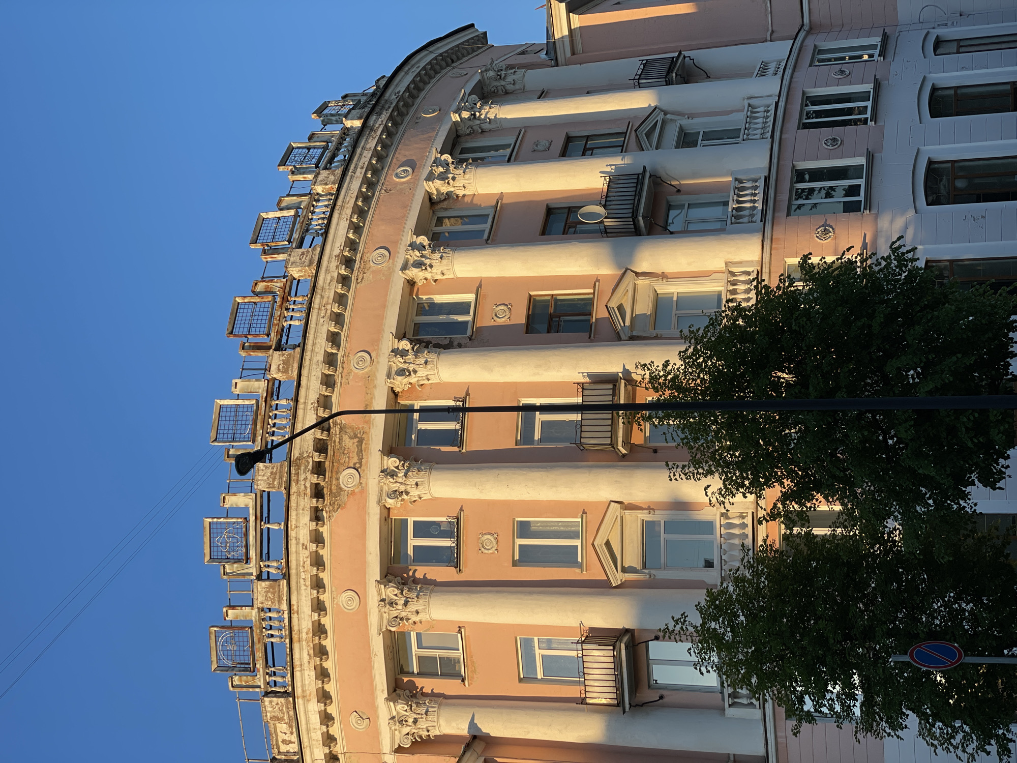 A low-angle, upward-facing view captures the facade of a curved building with architectural details like columns, ornate capitals, and projecting window cages. The building's exterior is primarily pink, with white architectural accents. Windows are seen with some open and some closed. Shadows cast by the setting sun highlight the building's curvature and the details of the columns. The sky above the building is a clear, bright blue. A tall, slender street light is visible in the foreground, partially obscured by green foliage from trees. A blue road sign is partially visible. This scene is taken at Zheleznogorsk, Russia.