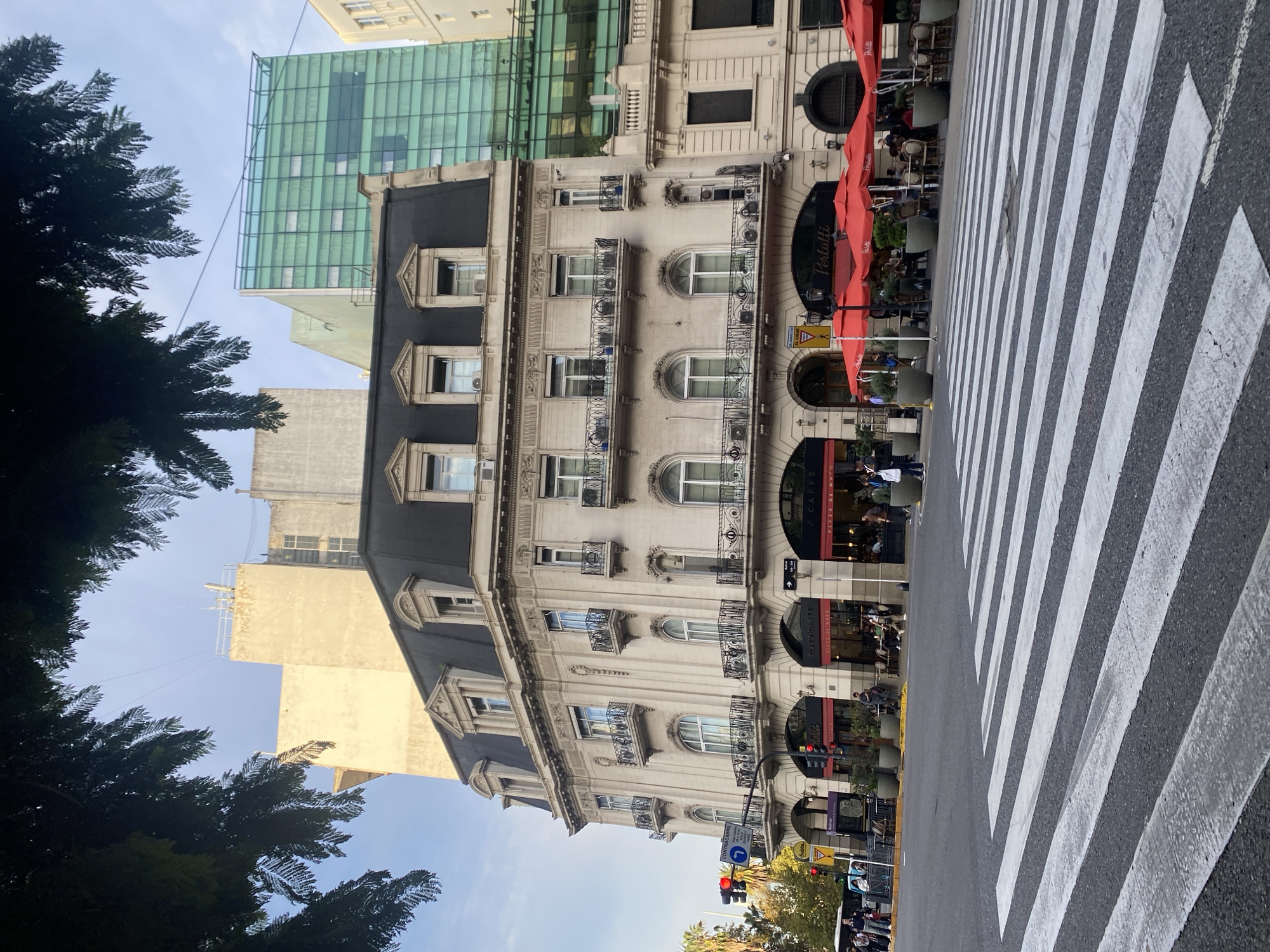 The image depicts a street scene in Buenos Aires, Argentina, looking up at an ornate building on the west side of Avenida Corrientes. The building is light-colored with dark-slated mansard roofs. It has multiple floors with arched windows and iron balconies, with "Plaza de Mayo" and "ristorante" signs. Red awnings cover outdoor seating. A modern green glass building stands behind it. Street level details include traffic lights, street signs, and numerous pedestrians. A crosswalk is visible in the lower right of the image, and trees are on the left side.