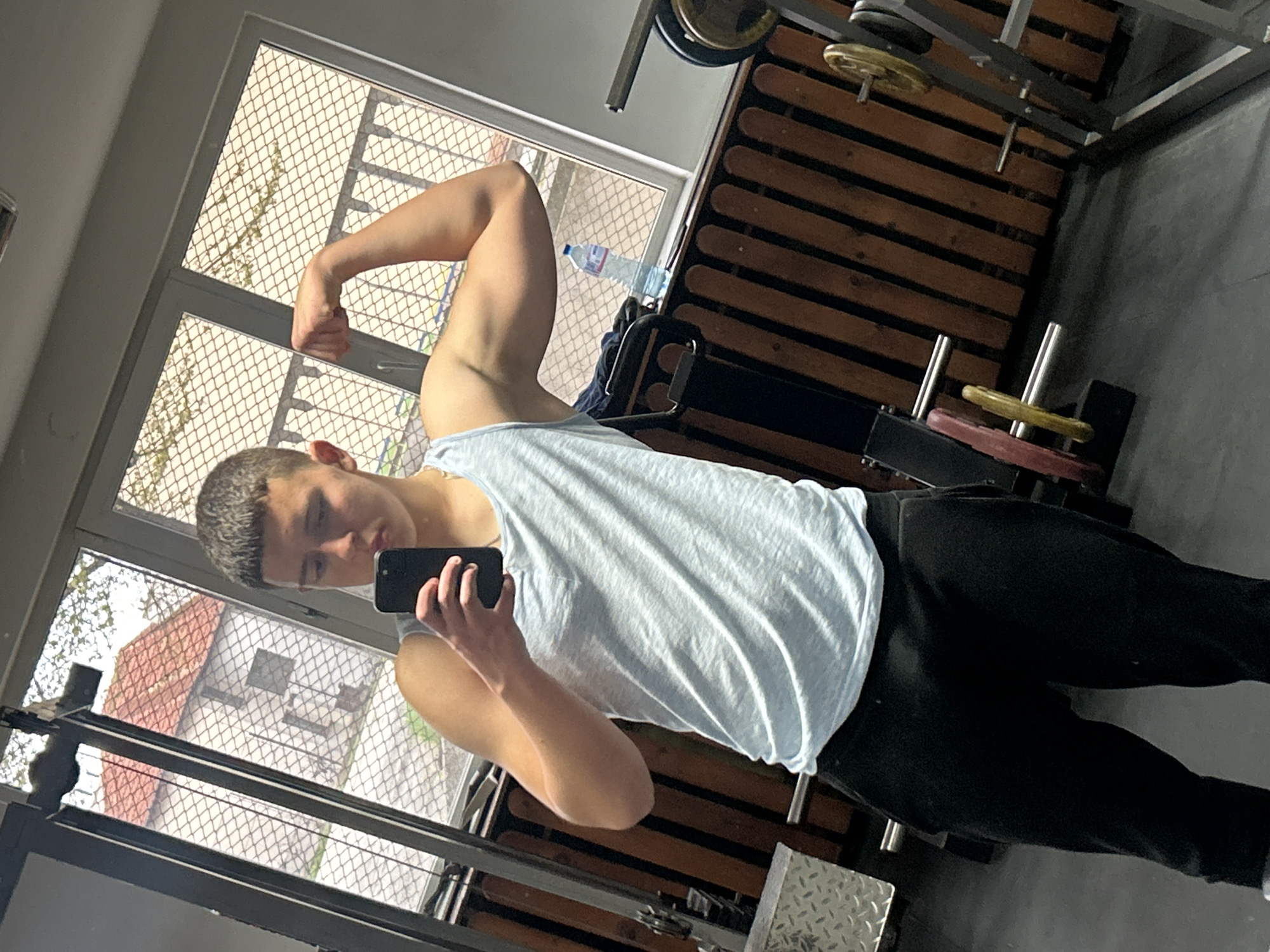 A young male is depicted in a mirror selfie, posing and flexing his right bicep, suggesting a moment of fitness documentation. He has short, dark hair and is dressed in a light blue sleeveless tank top and dark pants. His left hand is holding a black smartphone, indicating he is taking the photograph.
The setting appears to be an indoor gym or a dedicated workout area. Various gym equipment is visible, including weight plates in yellow, red, and black, stored on metal racks or bars. Portions of metal frames from exercise machines and a padded bench are also present. On the floor, a textured metal plate, likely part of gym equipment or flooring, is noticeable. A clear plastic water bottle with a blue cap is partially visible, resting on a piece of equipment near a window.
Through a large window, which is covered with a security mesh, an outdoor scene can be glimpsed. This includes the roof of a building with reddish-brown tiles and some foliage, providing a contextual clue for the location, which is Zlatograd, Bulgaria. The natural light filtering through the window suggests it is daytime. The subject's expression is neutral, focused on the photo-taking process, conveying a sense of dedication to physical fitness. No legible text is visible on any items in the image.