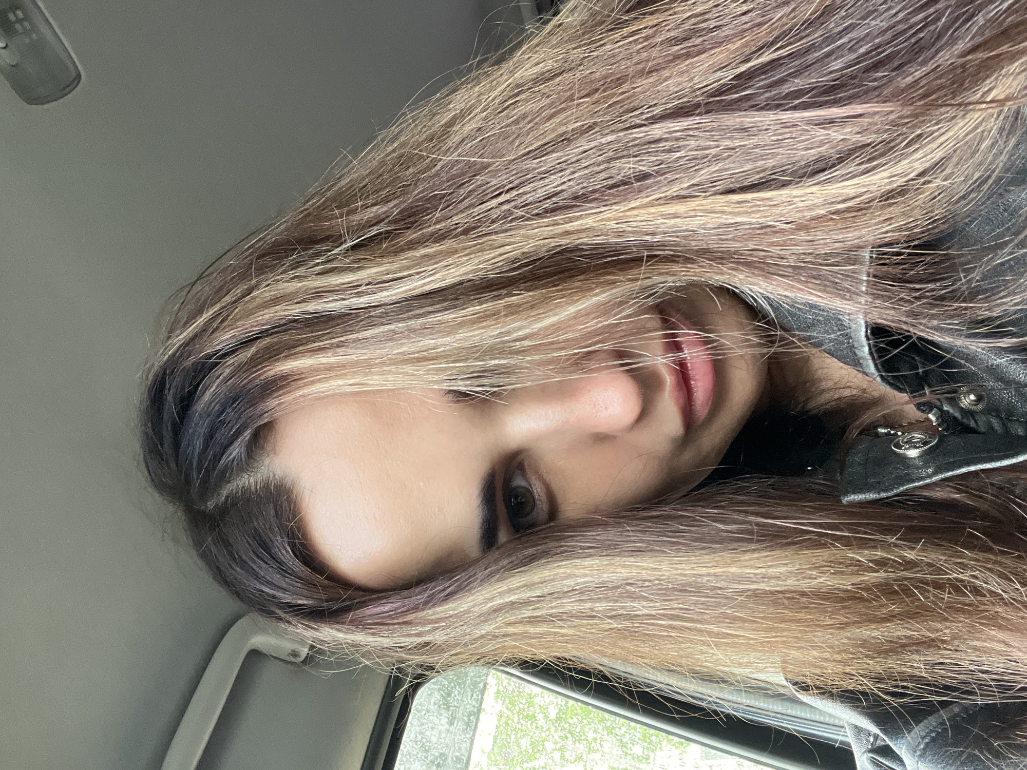 A close-up, vertically oriented portrait shows a person, likely female, inside a vehicle. The subject's head is tilted, with long, multi-toned hair flowing across the frame. The hair features dark roots blending into lighter shades of blonde and brown highlights, with hints of reddish or purplish tones visible in some strands.
Only part of the person's face is visible: one dark eye looking directly at the viewer, a portion of the nose, and slightly parted lips with a natural sheen. She has defined eyebrows. She is wearing a dark, possibly black, leather-like jacket with metallic buttons or studs and a small silver-colored charm near the collar.
The background indicates a car interior: a grey ceiling with a light fixture or sensor is visible above, and a grey grab handle is to the side. A window on the left side of the frame shows blurry green foliage and raindrops, suggesting it may be raining or overcast outside. No text is visible in the image.