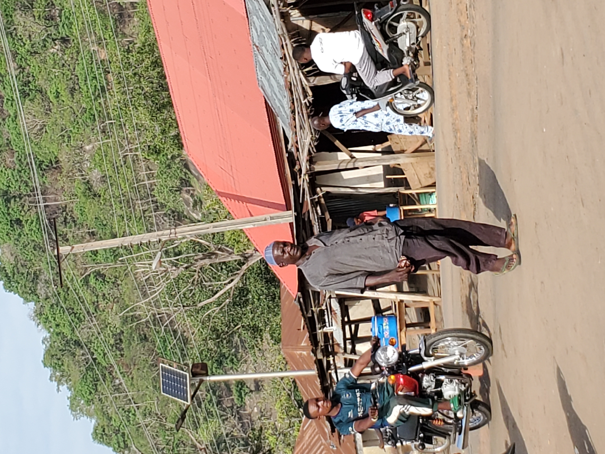 This image captures a vibrant street scene in Mararaba, Nigeria, under bright, clear daylight. In the foreground, a man wearing a green/blue patterned shirt and dark pants is seated on a red motorcycle, facing the viewer's left. Next to him, a utility pole is fitted with a small solar panel and a light fixture, suggesting a solar-powered street light.
In the middle ground, another man, dressed in a grey long-sleeved shirt, dark trousers, and a light blue cap, stands on the unpaved road, looking forward. Behind him and to his left, makeshift wooden stalls are set up beneath the eaves of a building with a striking red corrugated iron roof, which dominates the left side of the frame. Further back near these stalls, two other men are visible: one in a white shirt and grey pants stands beside a silver-colored motorcycle or scooter, and another wears a white and blue patterned shirt.
The background features a steep hillside densely covered with lush green vegetation and trees, suggesting a verdant natural landscape surrounding the settlement. Numerous overhead utility wires traverse the scene against the pale sky. The strong sunlight casts distinct shadows on the ground, indicating a sunny day. The scene depicts everyday activity in what appears to be a semi-urban or market area. No discernible text is visible.