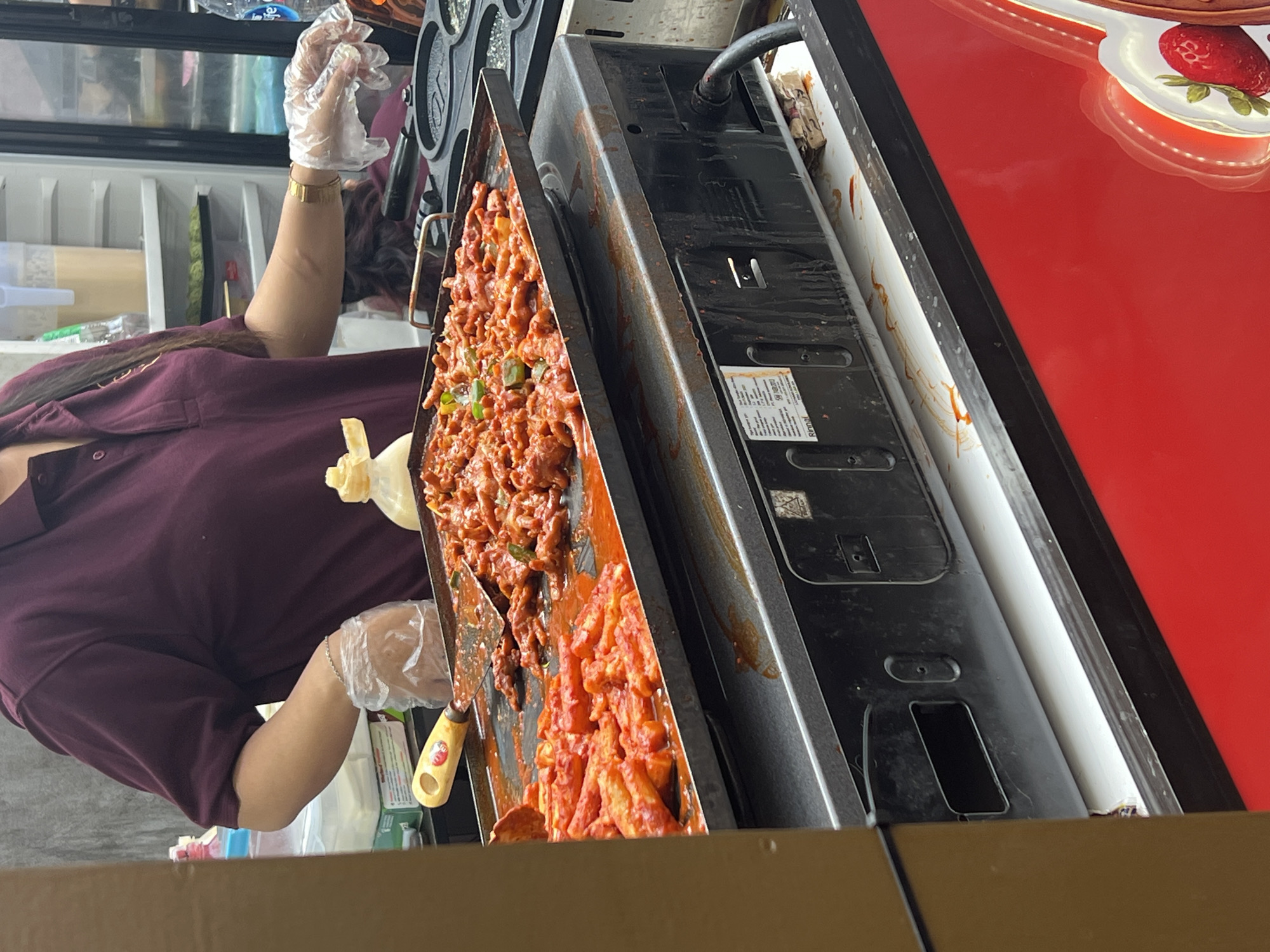 A vendor, wearing a maroon polo shirt and clear plastic gloves, is preparing food on a flat-top griddle. The griddle contains two distinct dishes: one filled with red, tube-shaped items in a sauce, and another with small, reddish, claw-like items, possibly chicken feet, also in a sauce. A spatula with a wooden handle rests on the griddle. The griddle is situated on top of a black appliance, possibly a stove or a food warmer, with visible vents and a power cord. The background shows shelves with containers and bottles, suggesting a food stall or market setting. The foreground features a bright red surface, possibly part of the stall's structure, adorned with a glowing image of strawberries. The scene likely takes place in a food stall in the city of Bandung, Indonesia, given the context provided.