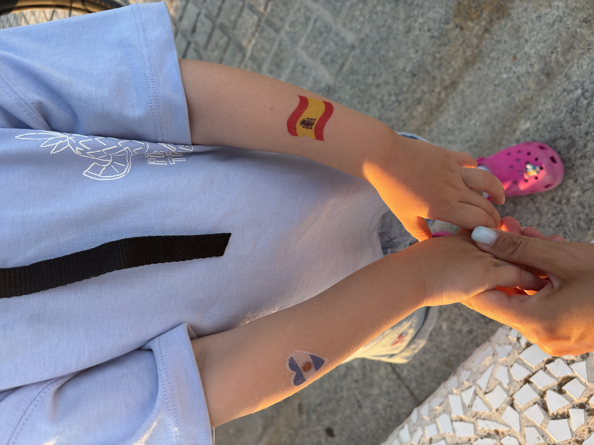 The image shows a close-up of a child's arms, adorned with temporary tattoos of national flags. On the left arm, a heart-shaped tattoo features the flag of Argentina. On the right arm, a wavy flag of Spain is depicted. The child is wearing a light blue t-shirt with a white graphic print and a black strap. The child's hands are clasped, one held by an adult, whose hand has light blue nail polish. The child is wearing pink Crocs. The background suggests an outdoor setting, possibly a park or promenade, with a stone-tiled bench on the left and paved ground. The lighting indicates it is either early morning or late afternoon, with warm sunlight casting shadows. The scene is likely in Valencia, Spain, given the Spanish flag tattoo.