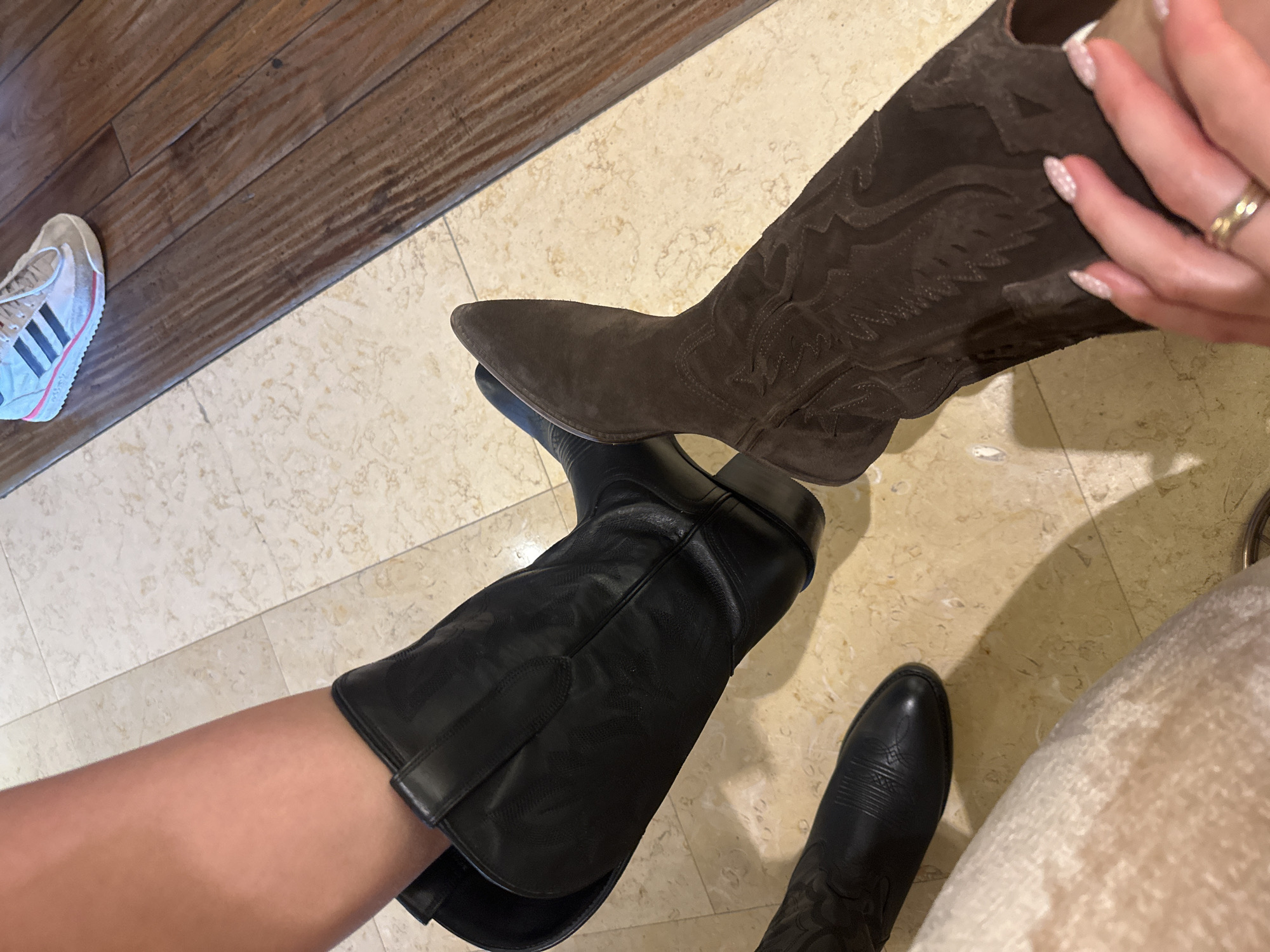A high-angle shot depicts two people wearing different pairs of cowboy boots. One person wears black cowboy boots, while the other wears brown suede cowboy boots. A partially visible Adidas sneaker lies on the left side. The scene appears to be indoors, set on a tiled floor. A wooden surface is also visible.