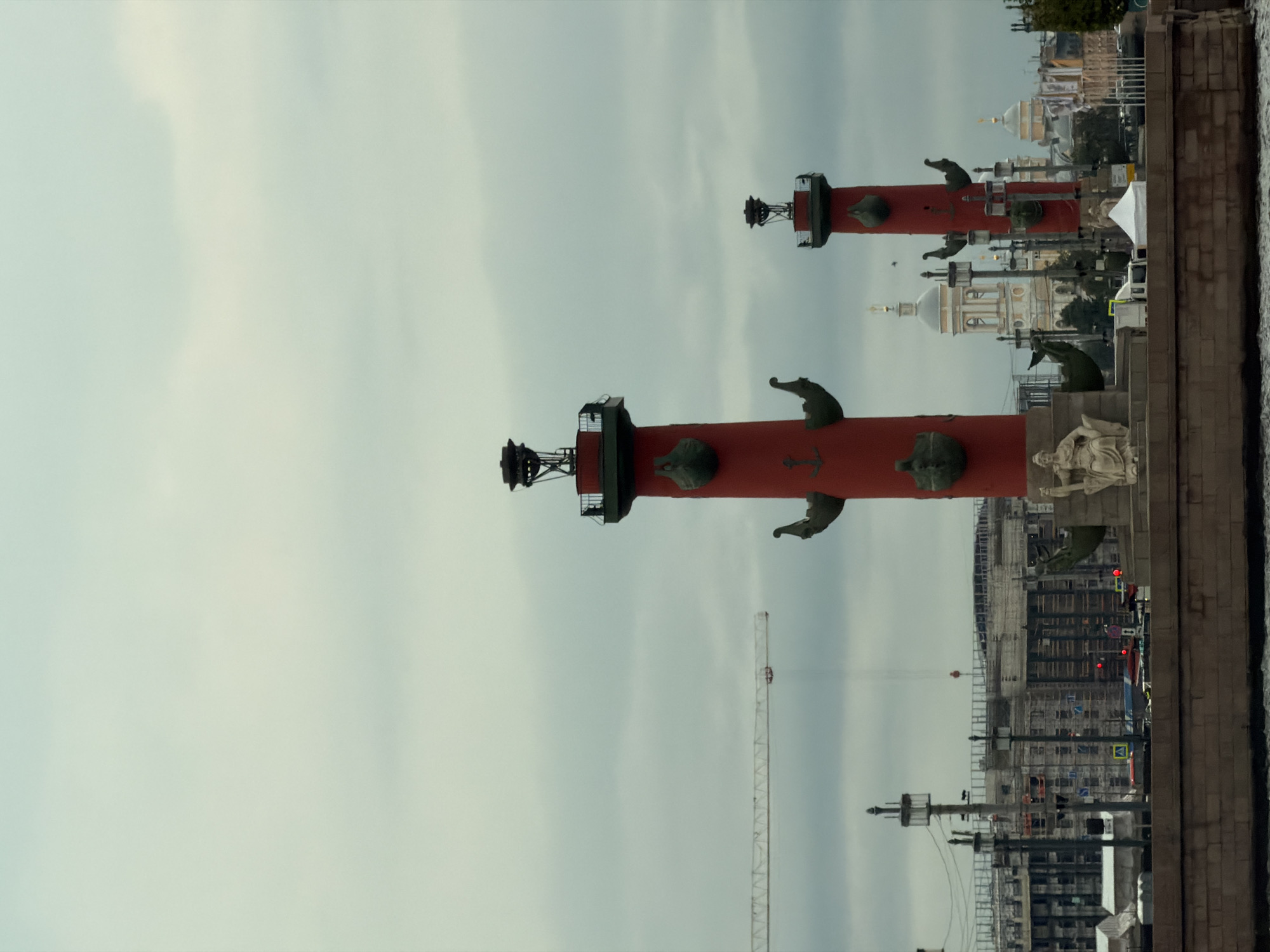 This image depicts a cloudy, overcast day in an urban waterfront environment. Dominating the foreground are two tall, red Rostral Columns, prominent landmarks that serve as former lighthouses. The columns are decorated with ship prows and anchors, and a statue of a seated woman is visible at the base of the left column.
Behind the columns, the cityscape unfolds. A large construction site is evident, with a long crane extending across the frame and numerous buildings under various stages of construction, some with scaffolding. In the distance, several domes of religious buildings are visible, including a large pale yellow dome with a cross on top. The water in the foreground appears calm, reflecting the muted tones of the sky. The overall atmosphere is somber and subdued, due to the cloudy weather. The location is Saint Petersburg, Russia, specifically on Vasilievsky Island, which is known for these Rostral Columns.