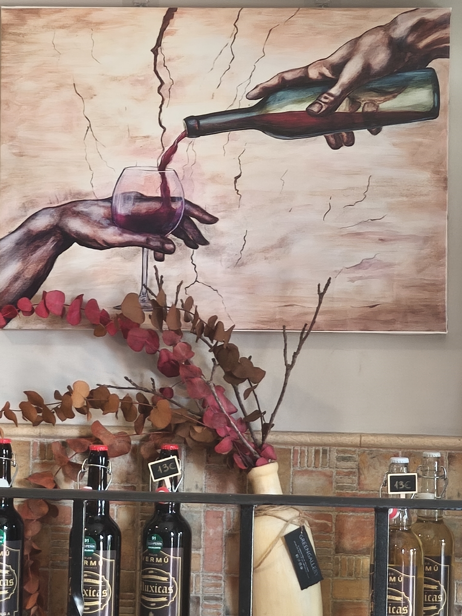 This image captures an indoor scene in Pontevedra, Spain, featuring a striking painting above a display shelf. The painting, rendered in warm, earthy tones with a cracked texture background, depicts a hand holding a dark-colored wine bottle, pouring red liquid into a wine glass held by another hand. The artistic style echoes classical art, reminiscent of Michelangelo's "Creation of Adam," but reinterpreted with a focus on wine.
Below the painting, a decorative arrangement of dried eucalyptus branches with vibrant red and brown leaves is placed in a light-colored, rustic jug or vase. The vase has a black tag tied with twine, partially revealing the text "GREENGALLERY".
The lower part of the image shows a black metal shelf displaying several bottles. Most are dark glass bottles with swing-top stoppers, labeled with text such as "VERMÚ" (Vermouth), "Muxicas", "91", and "PENIN". Some of these bottles have small black chalkboard price tags indicating "13€". To the right, lighter-colored bottles are also visible, one with a similar "13€" tag. The background wall behind the shelf features a textured, possibly tiled, surface in shades of brown and beige, resembling brickwork. The overall setting suggests a rustic yet inviting atmosphere, possibly in a wine shop, bar, or restaurant.