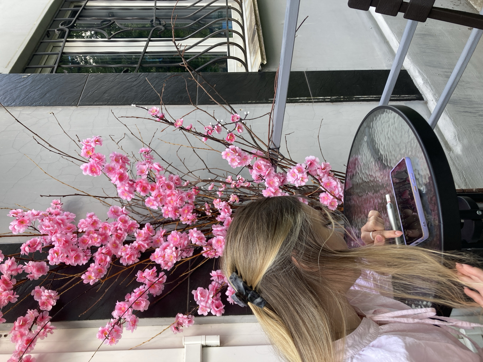 The media file captures a close-up view of a young woman with long, blonde hair as she styles it. She is seen from the back and side, with her hair pulled up and a dark hair clip securing it. Her right hand is actively working on her hair, while her left hand holds a silver stylus, possibly interacting with a phone or tablet. The phone, encased in a purple cover, is visible on a round, reflective glass table.
The scene is set outdoors, suggested by the natural lighting and the presence of decorative branches with pink artificial cherry blossoms. Behind the woman, a dark, textured wall or surface forms the backdrop for the floral display. To the upper left of the image, a window with an ornate metal grate is visible, hinting at a building's facade. The overall atmosphere appears casual and relaxed. There is no visible text in the image. The time of day seems to be daytime, as indicated by the bright, natural light.