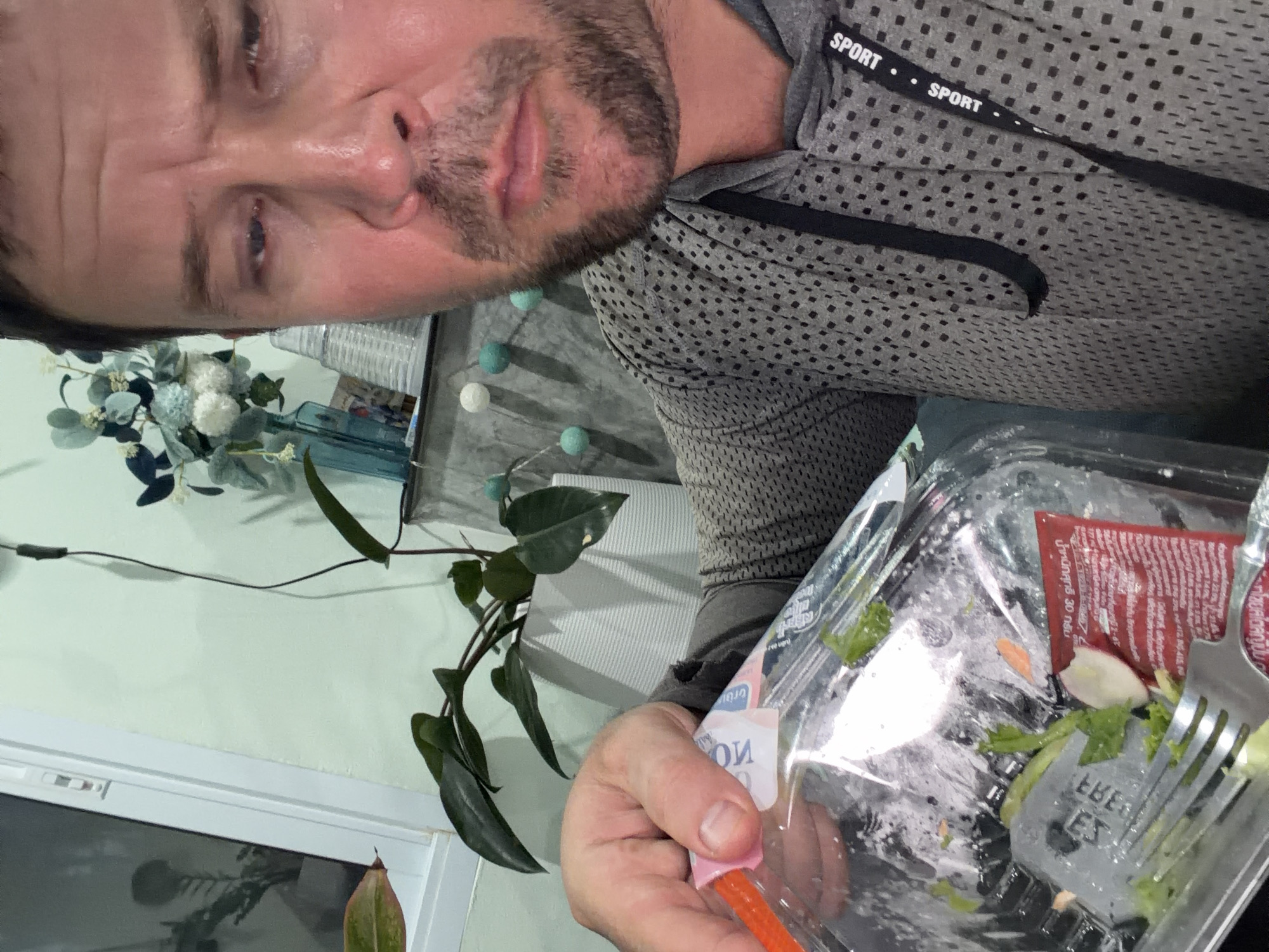 A man with fair skin, stubble, and sweaty skin is looking down with his eyes partially closed. He is holding a partially eaten salad in a clear plastic container with a fork inside. The salad appears to be mostly lettuce with some small pieces of tomato and radish. The man is wearing a grey hooded shirt with a black dotted pattern and a black lanyard with "SPORT" printed on it. In the background, there is a plant in a white pot, a window, and a shelf with decorative items. The scene appears to be indoors, possibly in a residential setting in Baan Mae Nam, Thailand.