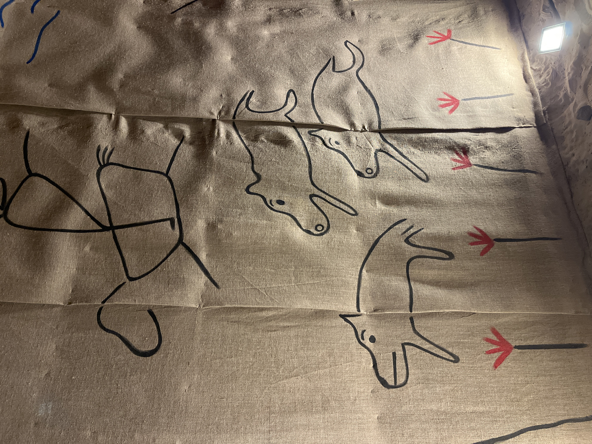 The image displays a vertical exhibit featuring primitive-style drawings on a creased, light-brown fabric, possibly linen or burlap. The fabric exhibits multiple prominent vertical folds and varying textures across its surface.
At the upper portion of the fabric, a large, stylized black outline depicts a human figure riding a quadruped animal, possibly a horse or deer, in motion towards the right. The rider is represented by basic stick-figure limbs and torso. Below this central figure, three smaller quadruped animals, resembling running dogs, wolves, or wild boars, are drawn in black outline, also moving towards the right. These animals have simple circular eyes and open-mouth indications. In the lower section of the fabric, four stylized plant-like forms are depicted with black stems and red, three-pronged tops.
The base of the display transitions into a textured, sandy-brown ground material. The scene is illuminated by an artificial light source, specifically a square white LED panel, which is visible at the bottom right, resting on the ground material and casting light upwards onto the fabric. This exhibit is consistent with a cultural or historical presentation found in a museum or similar institution within Saint Petersburg, Russia.
