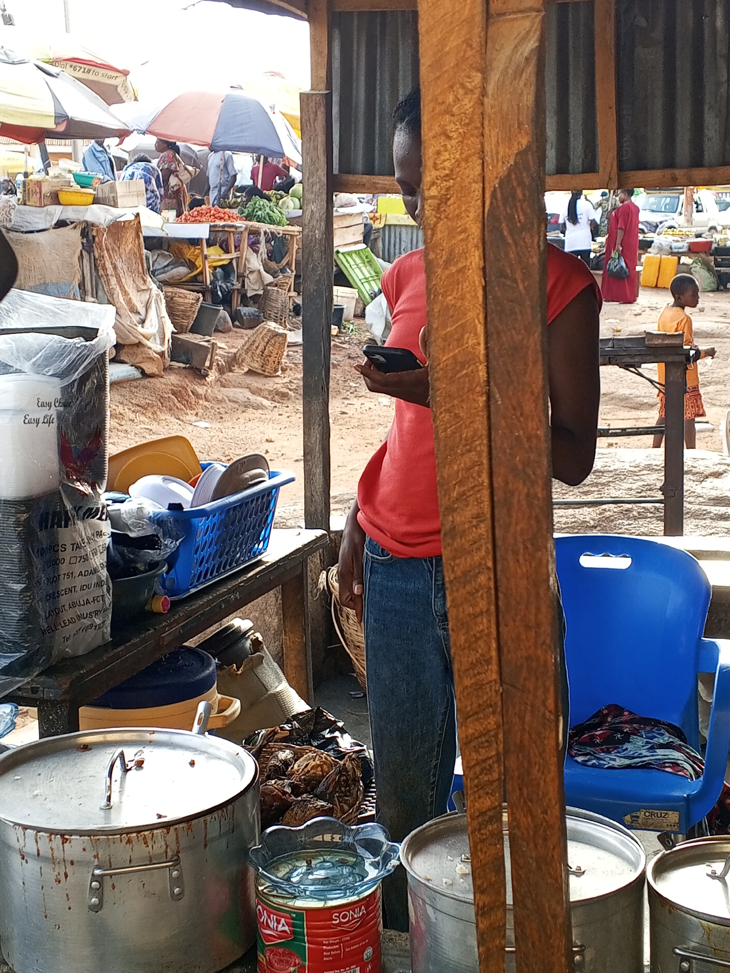 Here's a forensic description of the image:
**Location:** Gwong, Nigeria. An outdoor marketplace.
**People:**
* A person of African descent is standing holding a black smartphone in their left hand. They are wearing a short-sleeved, red t-shirt and blue jeans.
* Other individuals are visible in the background near fruit and vegetable stalls, and in other areas of the marketplace. One individual is wearing a red dress, and a child is visible near a table on the right.
**Subjects:**
* The primary subject is the person using the smartphone.
* Secondary subjects include market vendors, shoppers, and the general public visible in the background.
**Objects:**
* A plastic blue basket filled with dishes.
* Several large pots, one with the label "SONIA".
* Stacks of plastic disposable plates in packaging labelled "Easy Choice, Easy Life."
* A blue plastic chair.
* A wooden table surface.
* Market umbrellas and a variety of goods for sale.
**Actions:** The primary person is using their smartphone. Other people are moving about the market, presumably shopping and selling goods.
**Interactions:** The person is engaged with their phone. The other individuals appear to be conducting business and interacting within the marketplace.
**Environment:** The setting is a busy marketplace with stalls, tables, and various goods on display. The ground appears to be a red-brown color, possibly dirt or clay. Umbrellas are visible, suggesting sun protection or possibly rain protection. The scene is outdoors and suggests a relatively warm climate.