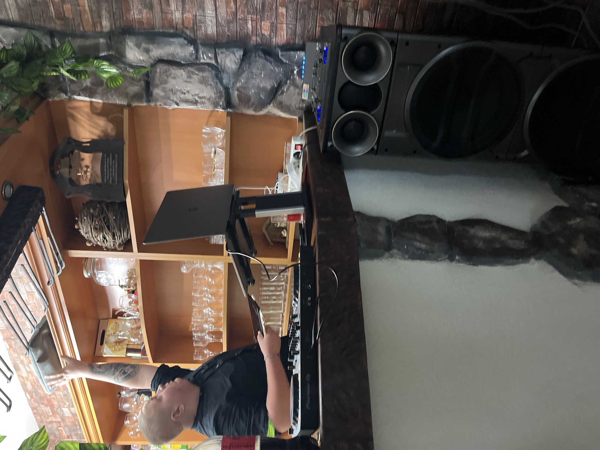 The image depicts an indoor scene, possibly in a bar or restaurant, within Presoka, Bulgaria.
A man with a bald head, wearing a black shirt and sporting tattoos on his arm, is standing at a bar or DJ setup. He appears to be operating the equipment. A laptop is positioned in front of him, and a Pioneer DJ controller is visible.
Behind the man, there's a wooden shelf unit filled with glassware and decorative items, including a woven basket and a metallic object resembling a sculpture.
To the right of the shelf, a large speaker system is positioned. Cables and power outlets are present nearby. The wall behind the shelf and speaker has a faux stone finish, and there's greenery visible in the upper left corner of the image. The environment suggests a setting catering to entertainment or social gatherings.