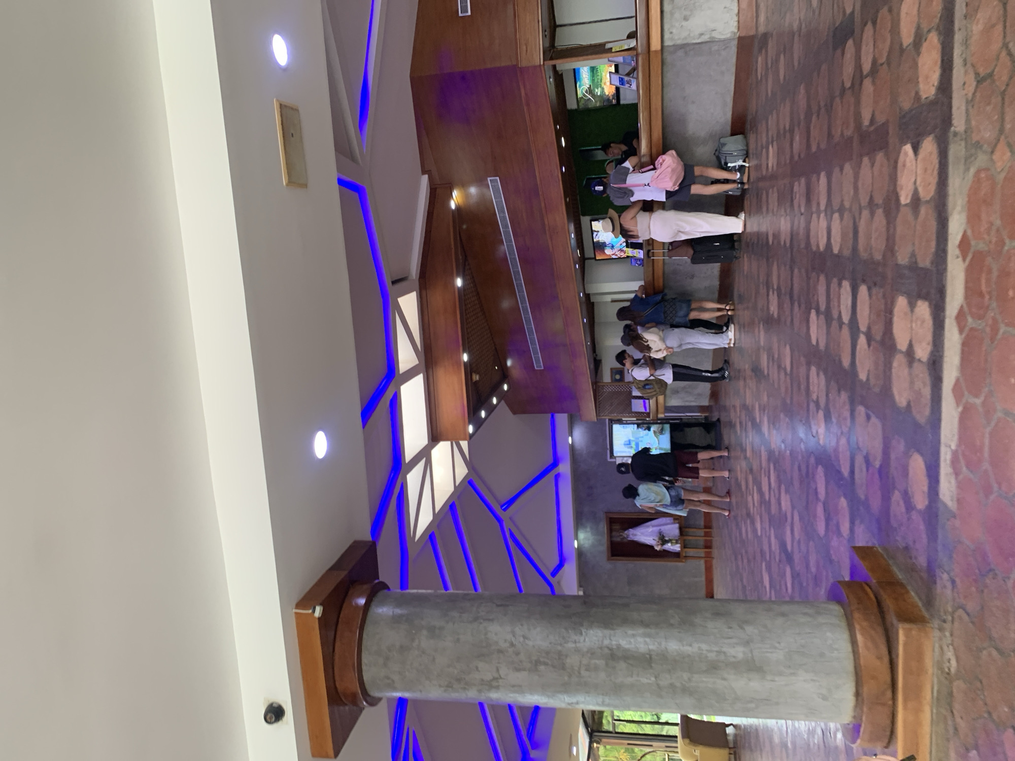 This image captures the interior of a hotel lobby or reception area located in Playa El Agua, Venezuela. The scene shows approximately ten individuals, primarily guests, queueing at a long reception desk made of wood with a textured grey lower facade. One female staff member is visible behind the counter, interacting with guests.
On the left side of the frame, a large cylindrical concrete column with a multi-tiered wooden capital and base stands prominently. To its left, a glimpse of an outdoor area or a brightly lit interior space with a yellow upholstered armchair is visible through a glass partition. Further back, against a grey textured wall, a white garment, possibly a wedding dress, is displayed on a stand, accompanied by a small floral arrangement on a wooden table.
The ceiling features a multi-level design: a flat white ceiling plane supports a lower, intricate structure above the reception area. This lower structure incorporates wooden panels and geometric patterns of bright blue LED strip lighting, along with recessed circular spotlights. Several horizontal silver air vents are integrated into the dark wooden paneling above the reception desk. Two security cameras are visible on the ceiling: one black camera in the upper left corner and a smaller white sensor/camera mounted on the wooden capital of the large column.
The floor is covered with reddish-brown octagonal tiles with darker grout lines, creating a distinct pattern. Guests at the reception desk are casually dressed, consistent with resort attire, including shorts, t-shirts, light pants, and dresses. Several individuals have luggage, including a black rolling suitcase, a grey duffel bag, and a pink tote bag, suggesting arrivals or departures. Two large screens behind the reception desk display text and imagery. The overall environment appears clean, well-lit, and designed for guest services.