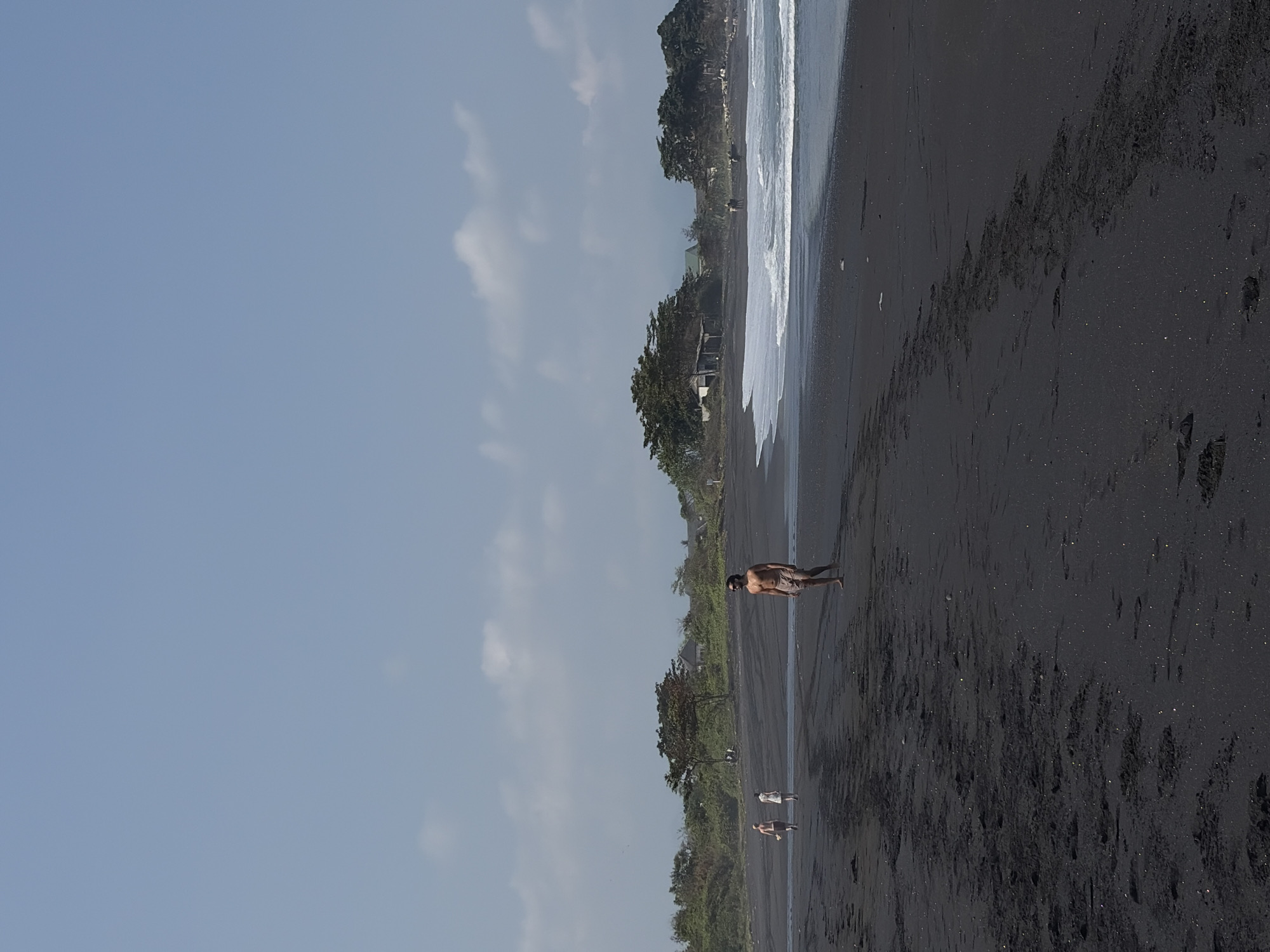 The image depicts a wide, black sand beach under a clear blue sky with sparse, white clouds. In the foreground, the dark sand is textured with footprints and small debris. A man, wearing light-colored shorts, walks towards the right of the frame, facing slightly away from the camera. Further down the beach, towards the left, two smaller figures, possibly children, are walking together. The middle ground of the image features a line of lush green trees and vegetation, with hints of buildings or structures nestled within. The ocean is visible on the right side of the frame, with gentle waves lapping at the shore, creating a white foam line. The scene appears to be tranquil and spacious.