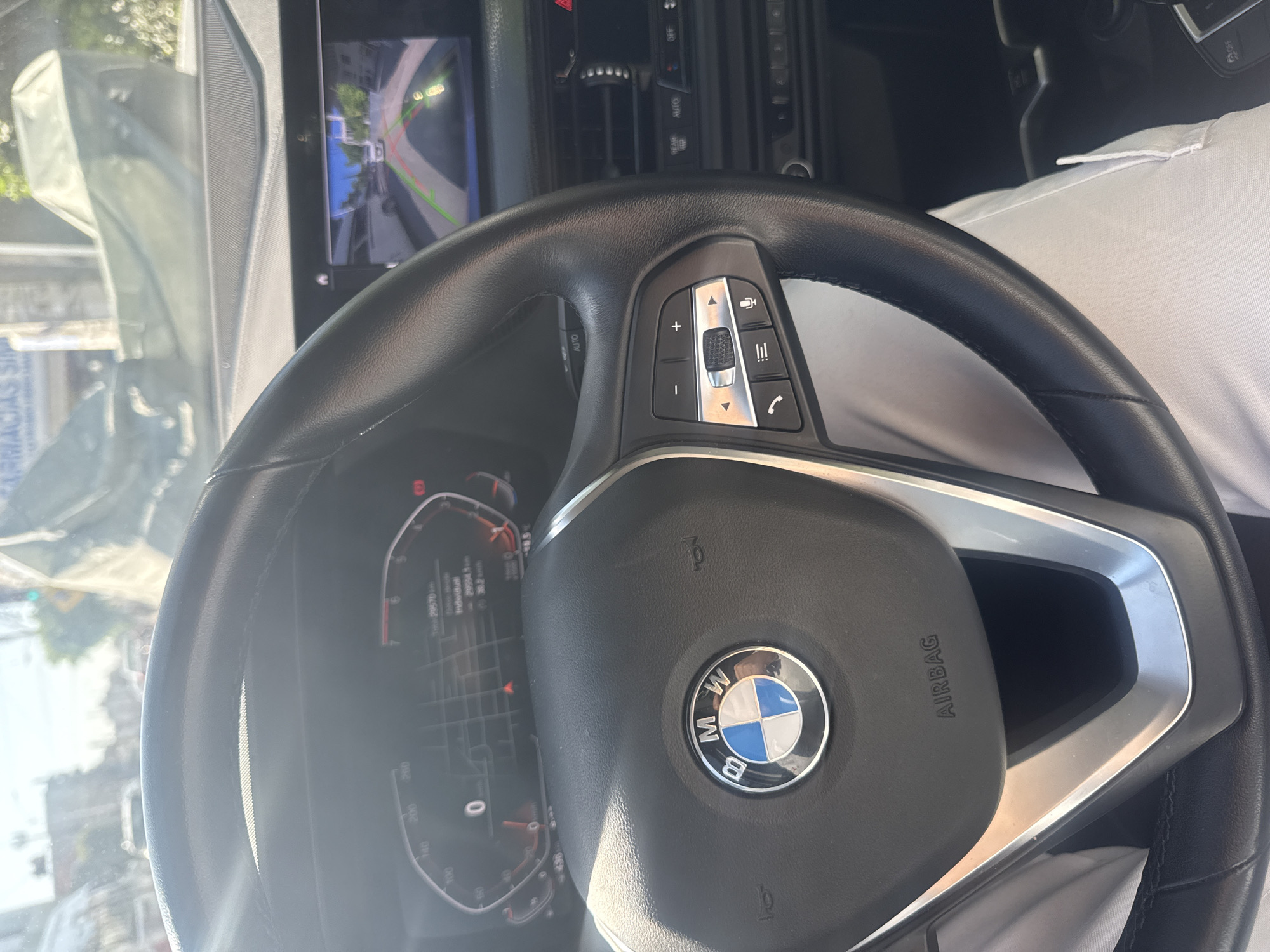 An eye-level shot inside a modern BMW car captures the steering wheel, dashboard, and a portion of the infotainment screen, all under bright daylight. A person, likely the driver, is visible in the passenger seat area, wearing a white short-sleeved collared shirt.
The black leather steering wheel, prominently featuring the blue and white BMW logo, is the central subject. To its right, a set of silver and black controls includes volume adjustment, track/menu navigation, phone call, voice command, and list/mode buttons, along with '+' and '-' buttons. The word "AIRBAG" is embossed on the steering wheel hub. To the left of the steering wheel, an "AUTO" button is visible.
Behind the steering wheel, the digital instrument cluster shows vehicle information, including a total mileage of "29570 km" and an individual trip mileage of "20954.1 km". A reading of "36.2" is also displayed, potentially indicating temperature. The speedometer shows markings like "0", "100", "140", "200", "260".
The infotainment screen, positioned above and to the right of the steering wheel, displays a reverse camera view, showing a street or alleyway with buildings, a curb, and parking lines. The camera view includes green and red guiding lines. Further into the dashboard, controls for "AUTO", "OFF", and "REAR TEMP" are visible.
Bright sunlight streams into the car, reflecting off the windshield and some interior surfaces, suggesting it is daytime with clear weather. Through the windshield, reflections and blurred elements outside the car are visible, including partial text that appears to be "FARMACIAS". The setting is likely an urban or residential area in Puebla, Mexico, given the location context. The car appears to be either parking or in reverse gear due to the active reverse camera.