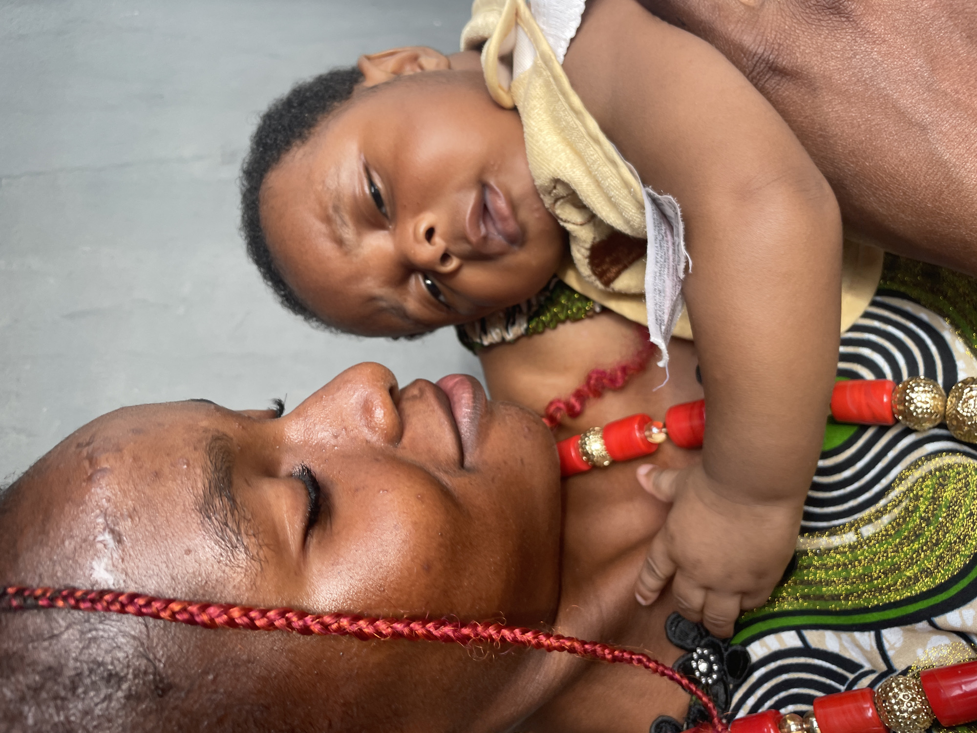 This vertical, close-up shot captures an intimate moment between a mother and her baby, set in Badagry, Nigeria. The mother, positioned at the bottom of the frame, has dark skin and her face is shown in profile, with her eyes closed, suggesting serenity or repose. A red braided adornment runs across her forehead. She wears a vibrant, patterned fabric dress featuring green, black, and white wavy stripes on a lighter background. Around her neck is a striking necklace composed of large, cylindrical red beads interspersed with smaller, ornate gold-colored beads.
The baby, also with dark skin, is held above the mother's chest, looking directly towards the camera with a curious and slightly open-mouthed expression. The baby wears a light yellow or beige top, possibly a bib or a simple garment, and a white fabric tag with illegible text is visible near its left arm. A small, dark green beaded necklace is also faintly visible around the baby's neck. The baby's hand gently rests on the mother's chest, near her necklace. The background is a plain, light grey, suggesting an indoor or sheltered setting. The image conveys a strong sense of warmth, love, and cultural pride, with the traditional attire adding to the local context.