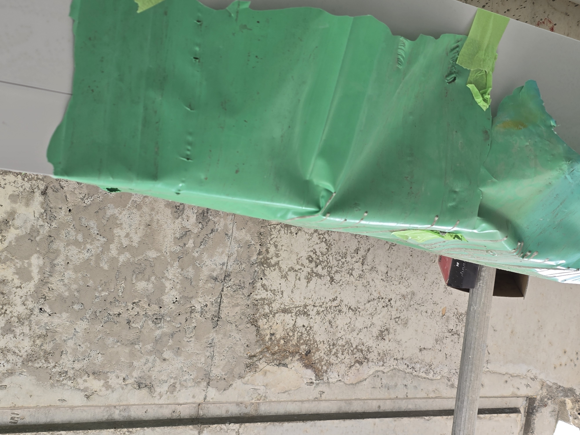 A medium, low-angle shot depicts a ceiling with a section covered in a wrinkled, semi-transparent green plastic sheet, secured in place with strips of green tape. The ceiling itself has a mottled, textured surface with areas of disrepair and staining. A metallic support pole runs vertically, supporting the structure. The location is Unknown, Unknown, but given the unfinished state, it appears to be a construction or renovation site. No people are visible in the scene.