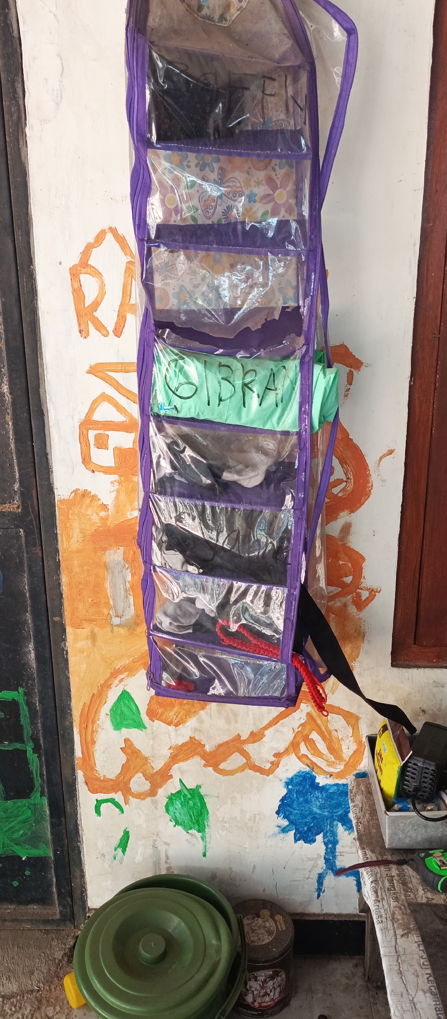 An exterior scene in Jatisari, Indonesia features a white wall with orange and green paint splatters, possibly from children's artwork. A hanging organizer with multiple clear plastic compartments, bordered in purple, is attached to the wall. Inside the organizer are various items, including what appear to be folded clothes. One compartment clearly displays the handwritten name "GIBRAN" in black marker on a light green background. To the left of the organizer, on the wall, is painted an orange house-like structure with a yellow roof. On the ground beneath the organizer, there is a green plastic bucket with a lid, a yellow toy, and two cylindrical containers. To the right of the organizer, a wooden shelf holds various objects including a yellow and black electrical device, a tape measure, and some paper. A dark wooden door frame is visible on the far right.