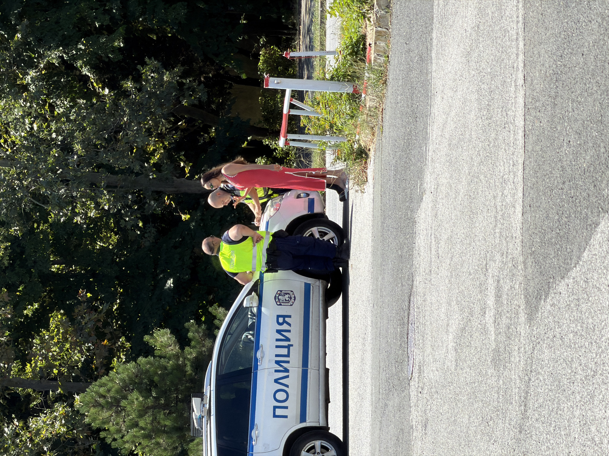 The image depicts a scene on a paved road in the city of Burgas, Bulgaria, with lush green trees and foliage in the background. A white police car, marked with blue stripes and the word "ПОЛИЦИЯ" (Police), is parked on the right side of the road. Two police officers are present. One officer, wearing a bright yellow safety vest over dark blue trousers, stands by the driver's side of the car, with his back to the camera. Another officer, also in a yellow vest and dark trousers, is interacting with a woman. This woman, dressed in a long, sleeveless red dress with white trim, is standing near the front of the police car, looking down at a document that the officer appears to be holding or presenting to her. The interaction seems formal, possibly involving an exchange of information or a traffic stop. A white and red barricade is visible in the background, near some overgrown vegetation. The overall atmosphere appears to be that of an official police interaction on a sunny day.