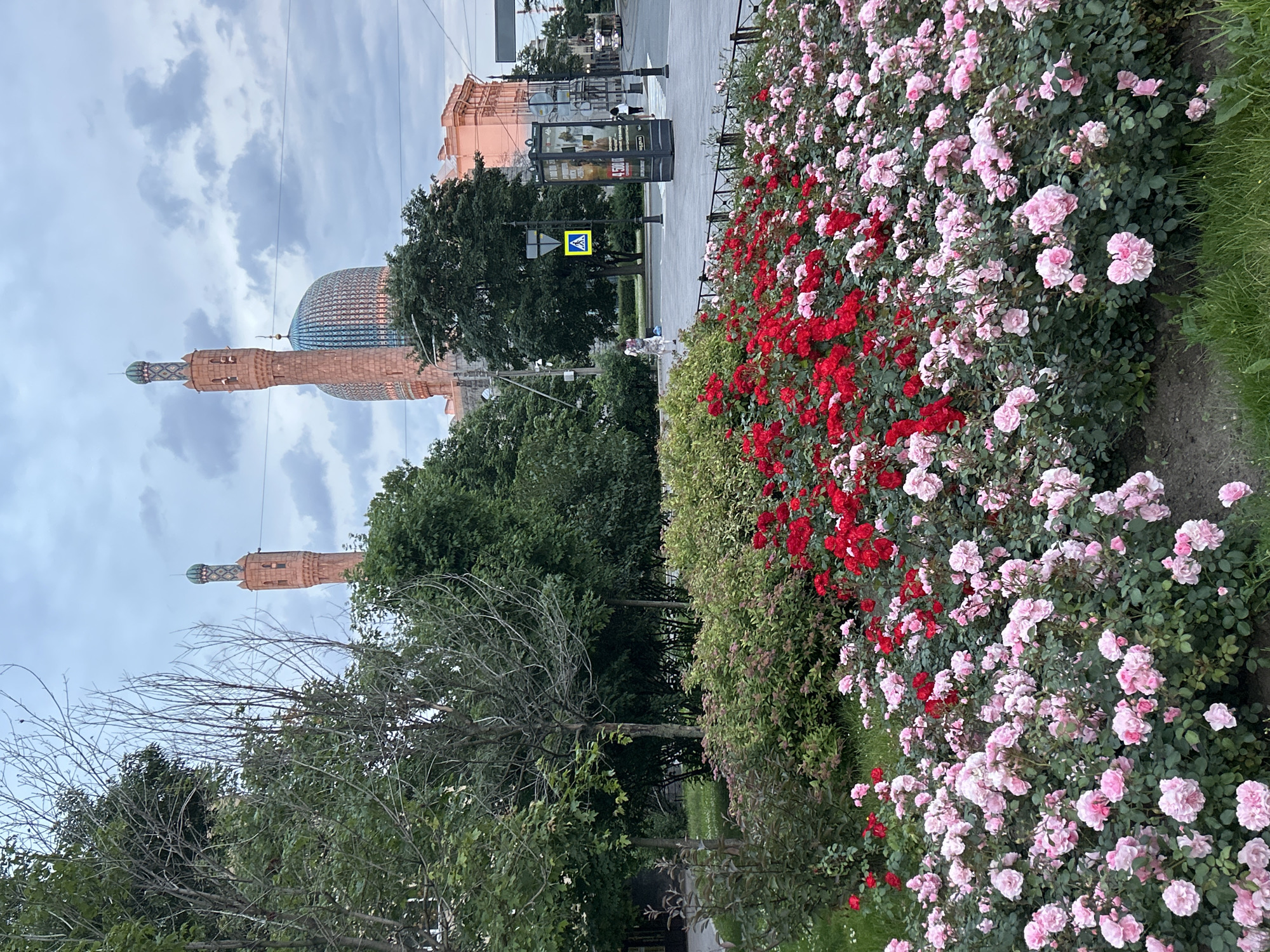 The image displays a vibrant flowerbed filled with an abundance of pink and red roses, taking up the majority of the right side of the frame. To the left, a street scene unfolds under an overcast sky. In the background, a large, ornate building with a distinctive dome and tall towers is visible, hinting at a historical or religious site. A pedestrian crossing with a yellow warning sign is also present, along with a streetlamp and what appears to be a bus stop or advertising kiosk. The presence of trees and the overall composition suggest an urban setting, possibly a park or a square adjacent to a significant landmark. The soft lighting and cloudy sky indicate it might be late afternoon or early morning, with a generally calm atmosphere. No explicit text is legible in the image.