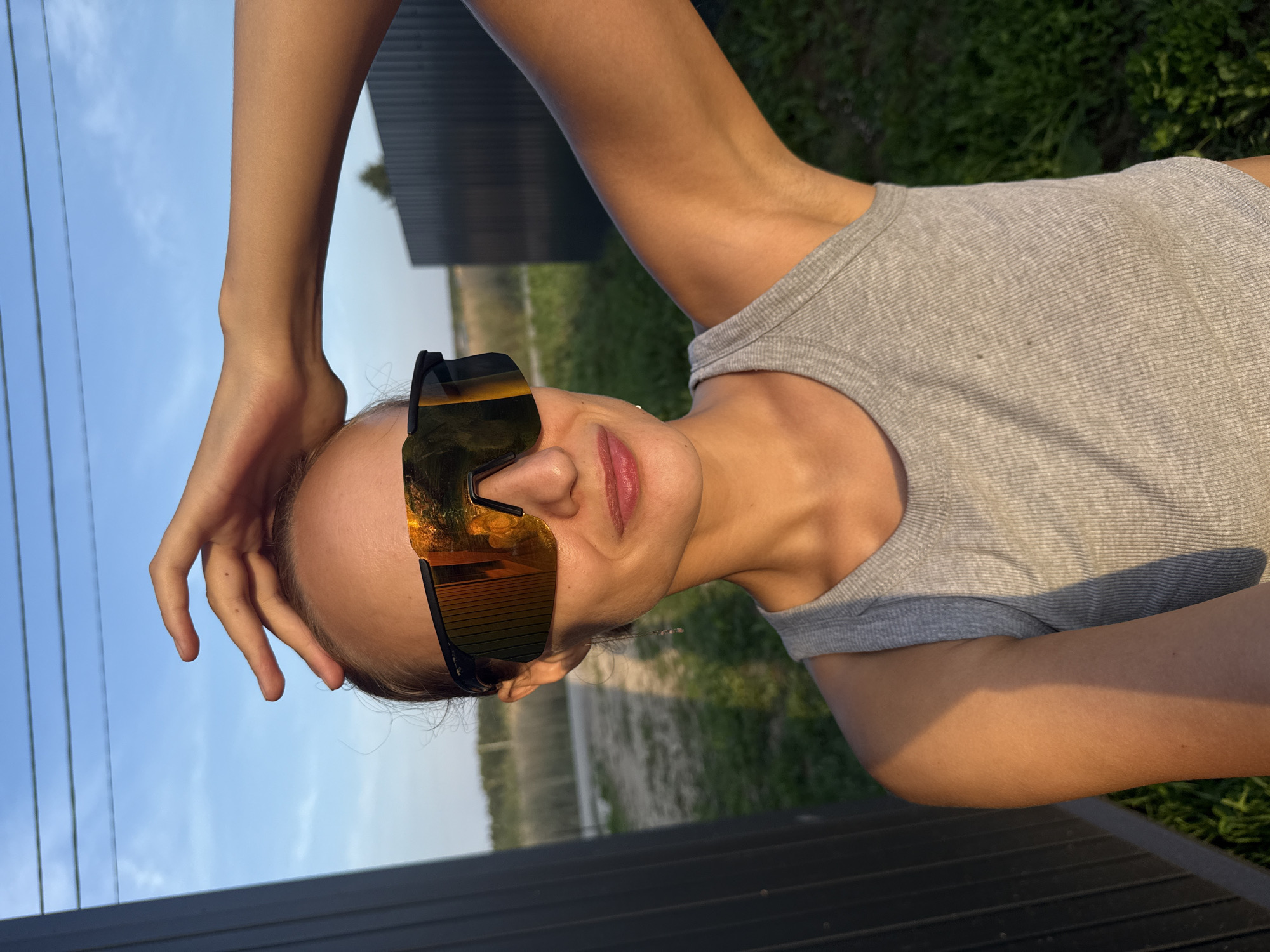 A young woman, wearing mirrored wraparound sunglasses and a grey ribbed tank top, is pictured outdoors. Her right hand is raised, resting on her head. The background features a clear blue sky with power lines, a dark fence on the left, and a grassy area with some vegetation. The lighting suggests it is late afternoon or early evening, with the sun casting a warm glow on the subject. The location is identified as Podshivalovo, Russia.