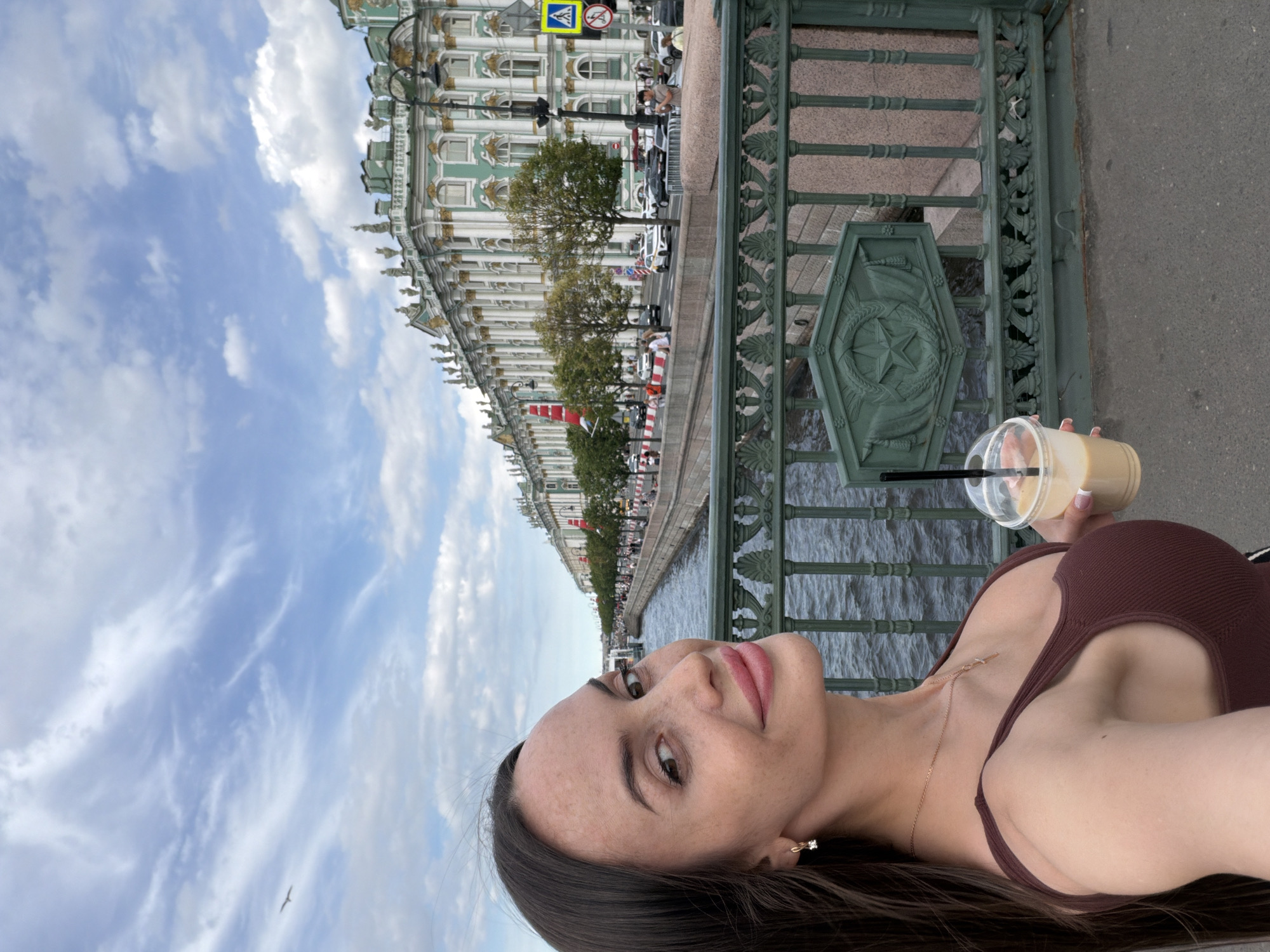 A woman with long dark hair is taking a selfie while standing on a bridge, holding a to-go cup with a beverage. She is wearing a brown tank top and a necklace. In the background, a canal or river flows alongside a grand, ornate building, likely the Winter Palace in Saint Petersburg, Russia. The building is characterized by its classical architecture, featuring numerous columns, statues, and green accents. Across the water, there is a street with trees, pedestrian traffic, and cars. The sky is partly cloudy, with wispy white clouds against a blue backdrop. The bridge railing in the foreground is made of dark green metal with decorative elements.