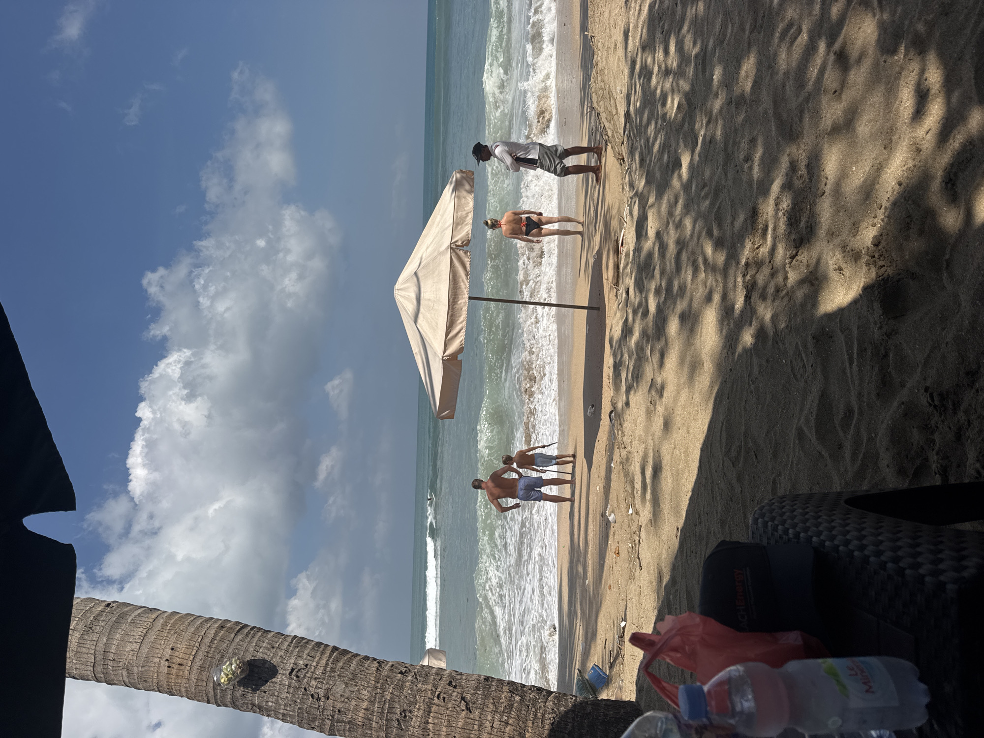 The image depicts a sunny day on Kuta Beach, Indonesia. In the foreground, a palm tree trunk stands on the left, with a glass object containing what appears to be small, round items attached to it. A beach chair with a black cap and a red bag rests in the lower left corner, partially in shadow. Beyond the chair, the sandy beach stretches towards the ocean.
In the middle ground, several people are present. A man and a boy stand near the water's edge, facing the sea. The man is wearing blue swim trunks, and the boy is in light-colored shorts. Further to the right, a woman in a black bikini stands under a beige beach umbrella. A man in a white shirt and grey shorts stands nearby, facing away from the viewer. The ocean is active with waves breaking on the shore, and a surfer is visible in the distance on the water. The sky is blue with scattered white clouds. The beach appears somewhat littered with small white objects and debris.