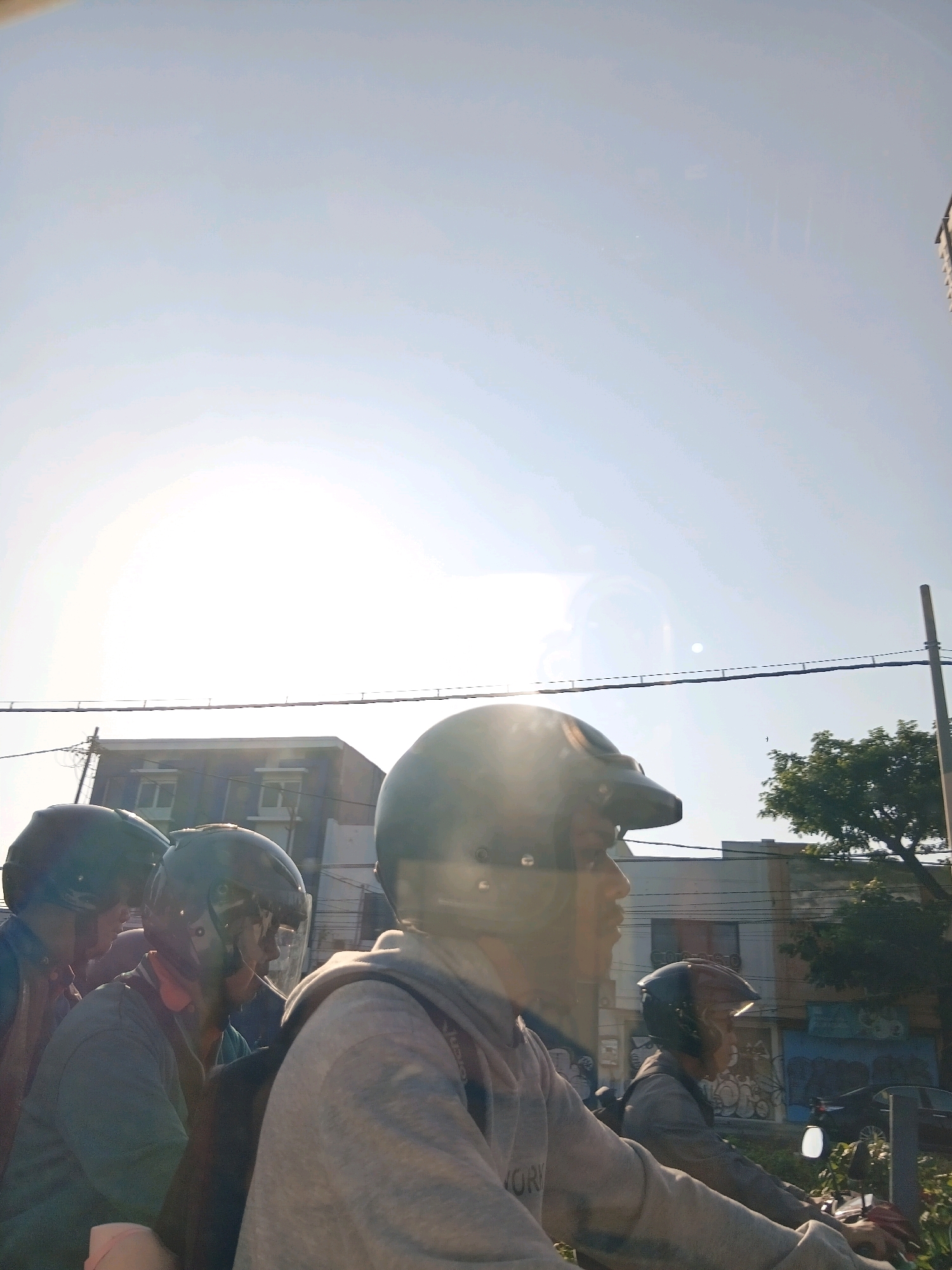 The image depicts several individuals on a motorcycle, viewed from the side, under a bright, hazy sky. The sky is the dominant element, taking up the majority of the frame and exhibiting a lens flare effect. In the background, a multi-story building with a dark facade and white-trimmed windows is visible. To the right of the building, a green tree stands out. A utility pole with a wire strung between two points is also present in the middle ground. The people are wearing helmets, and their clothing appears casual. The scene suggests an outdoor setting, likely a street or road, given the presence of vehicles and buildings. The hazy sky and lens flare suggest a sunny day, possibly with some atmospheric conditions affecting visibility. The overall impression is of a candid moment captured during transit.