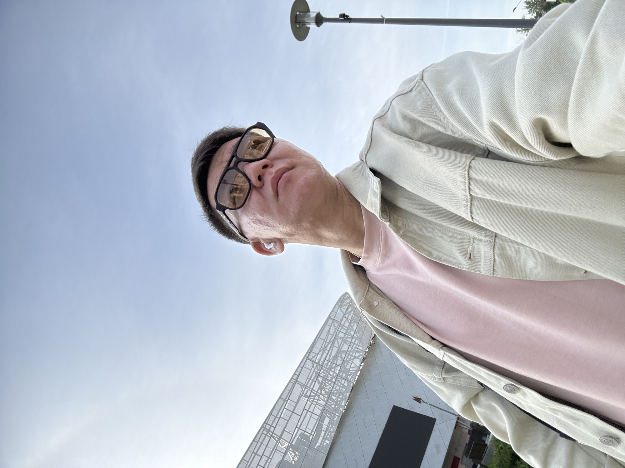 The image shows a man wearing glasses, a pink t-shirt, and a light-colored jacket. He is looking up at the camera. In the background, there is a blue sky, a lamp post, and a modern building with a grid-like facade. Based on the architectural style, the location is likely the VTB Arena in Moscow, Russia.