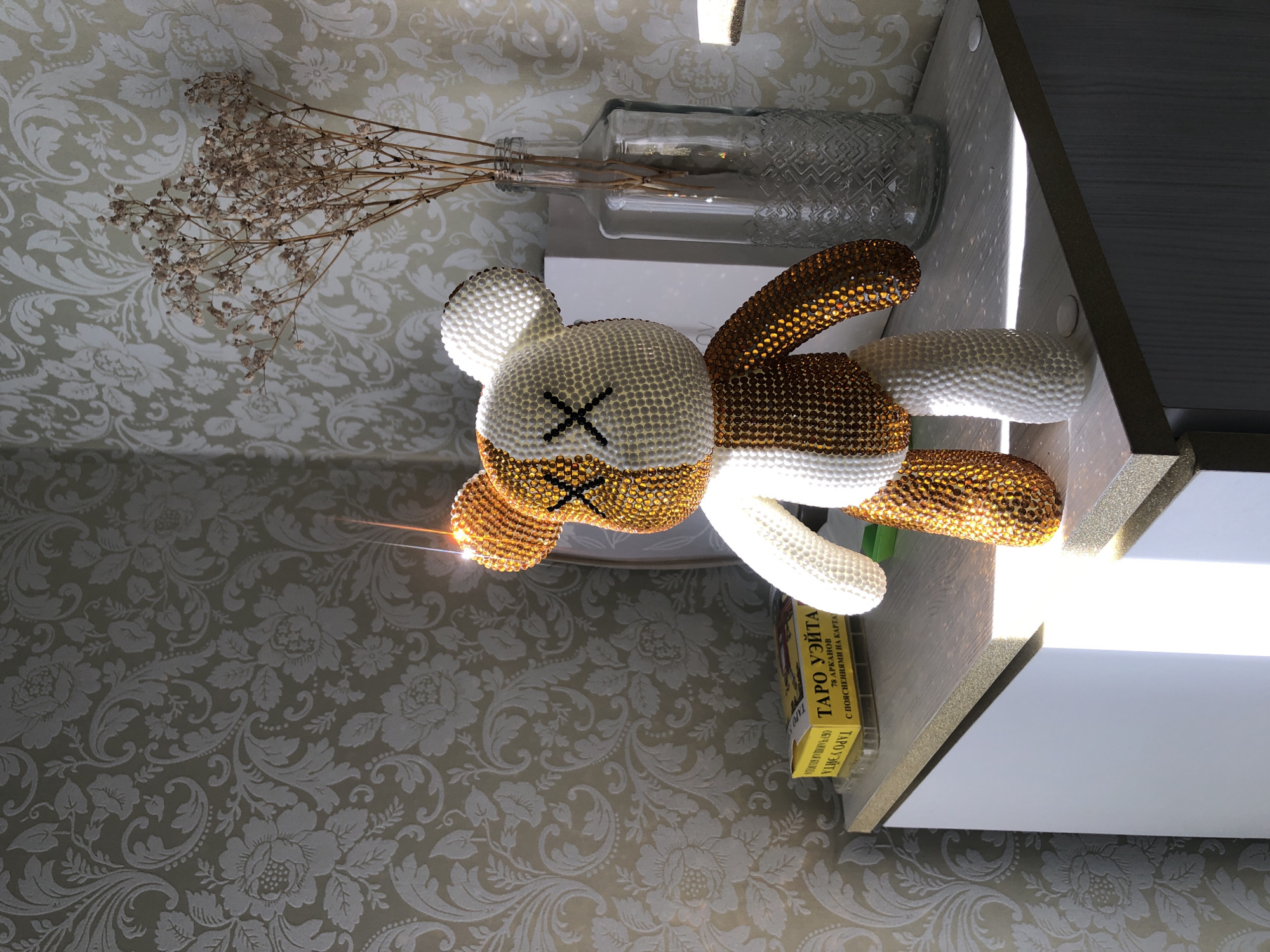 Here is a detailed description of the image:
The image depicts a close-up, slightly overhead shot of a decorative arrangement on what appears to be a shelf or tabletop. A beaded bear-like figure with "X" marks for eyes lies centered in the frame. The figure is white and gold-brown, with shiny beads covering its surface. Next to the figure is a glass vase holding dried flowers. A box of tarot cards sits to the right of the figure. The shelf or tabletop is positioned in front of a wall covered in patterned wallpaper. The scene is brightly lit, with a light source illuminating the shelf from the right side.
There is no indication of the location of the city of Noviy, Russia in the image.